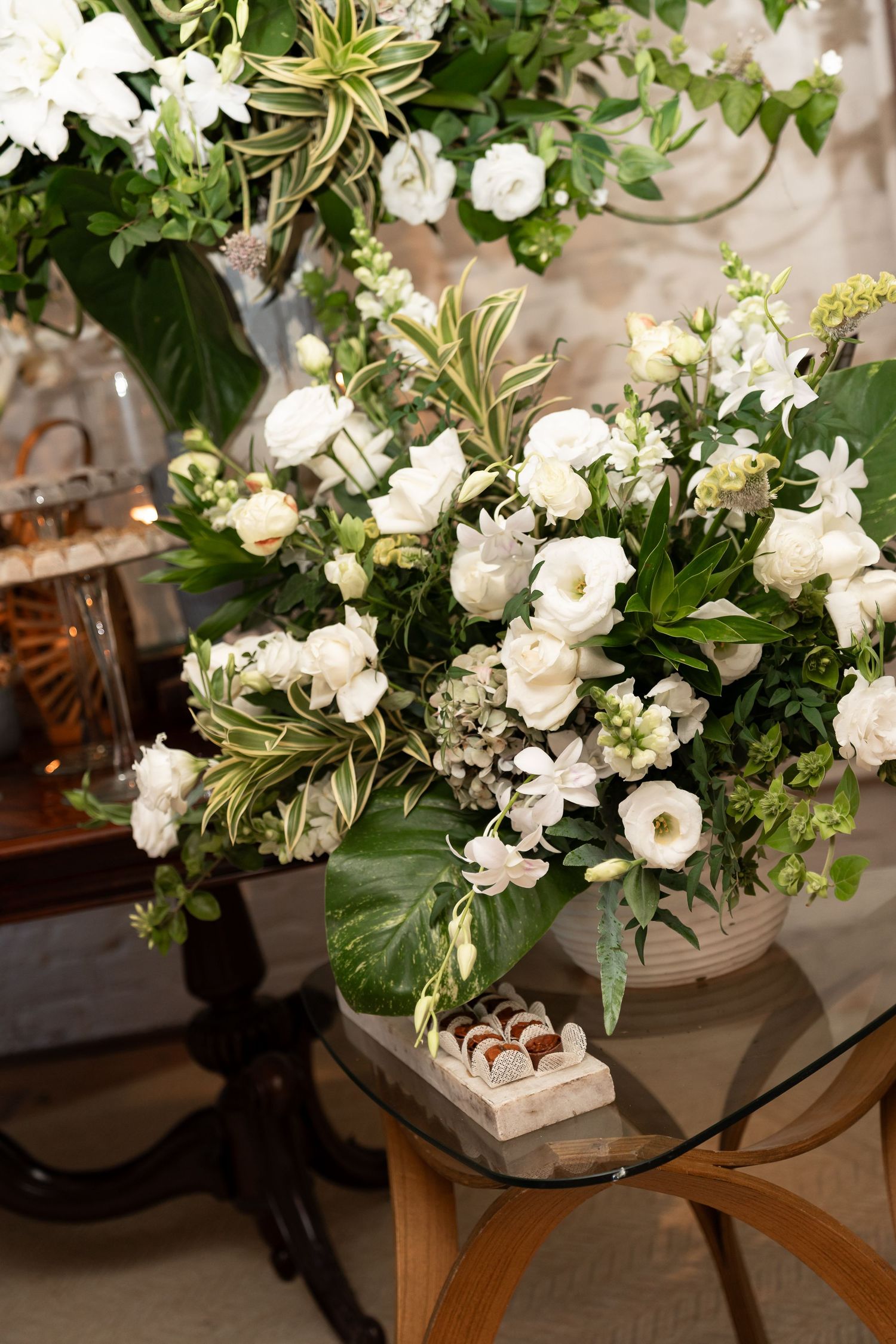 Elegant white floral arrangement with greenery in a ceramic vase on a glass table, with chocolates nearby.