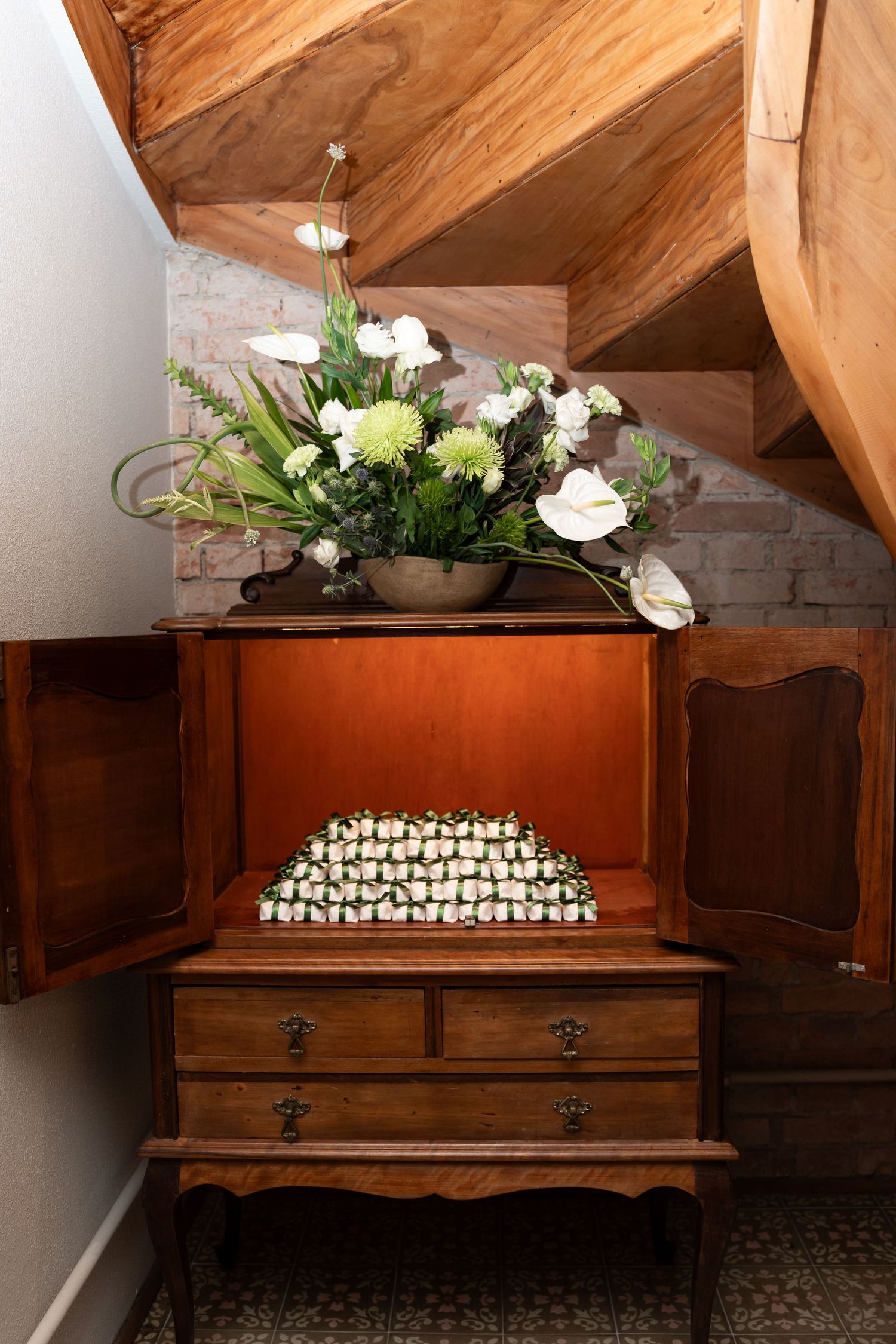 Antique wooden cabinet with open doors displaying white flowers inside, topped with a floral arrangement under wooden beams.