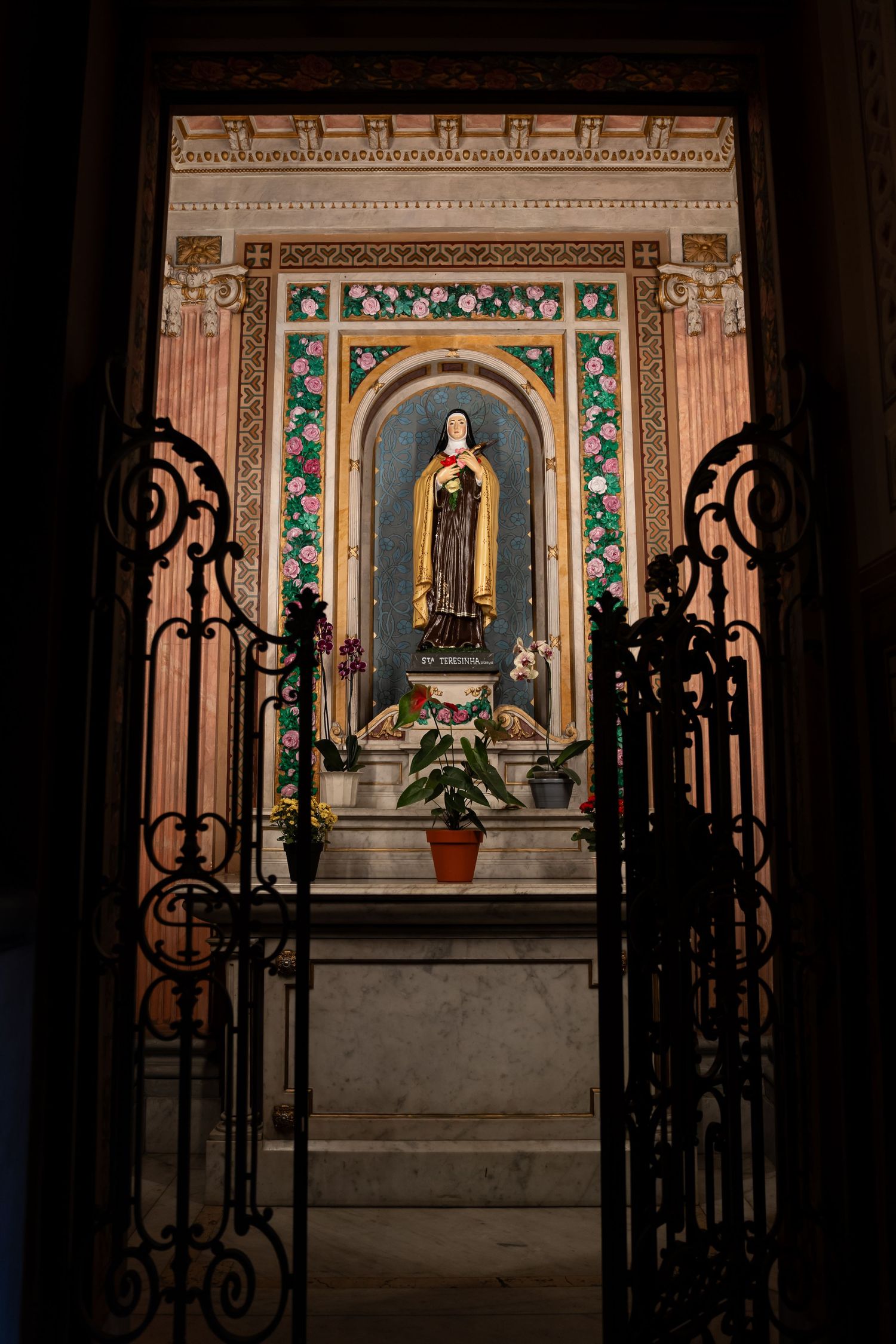 Ornate Catholic chapel altar with a saint statue in a blue niche, framed by floral decor and wrought iron gates.
