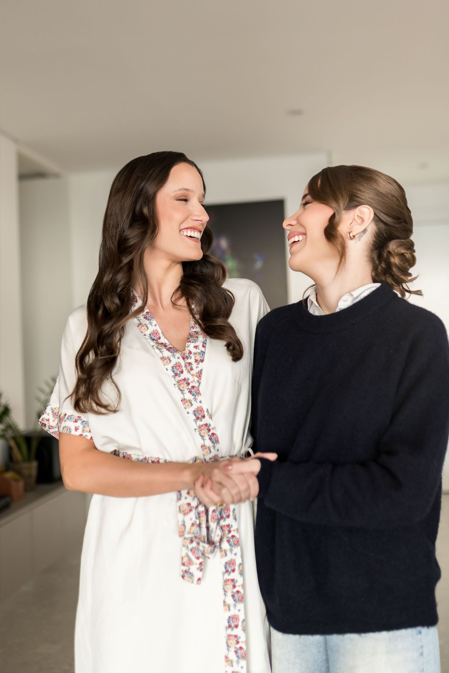Two smiling women holding hands and laughing together indoors, one in a white floral dress and one in a navy sweater.