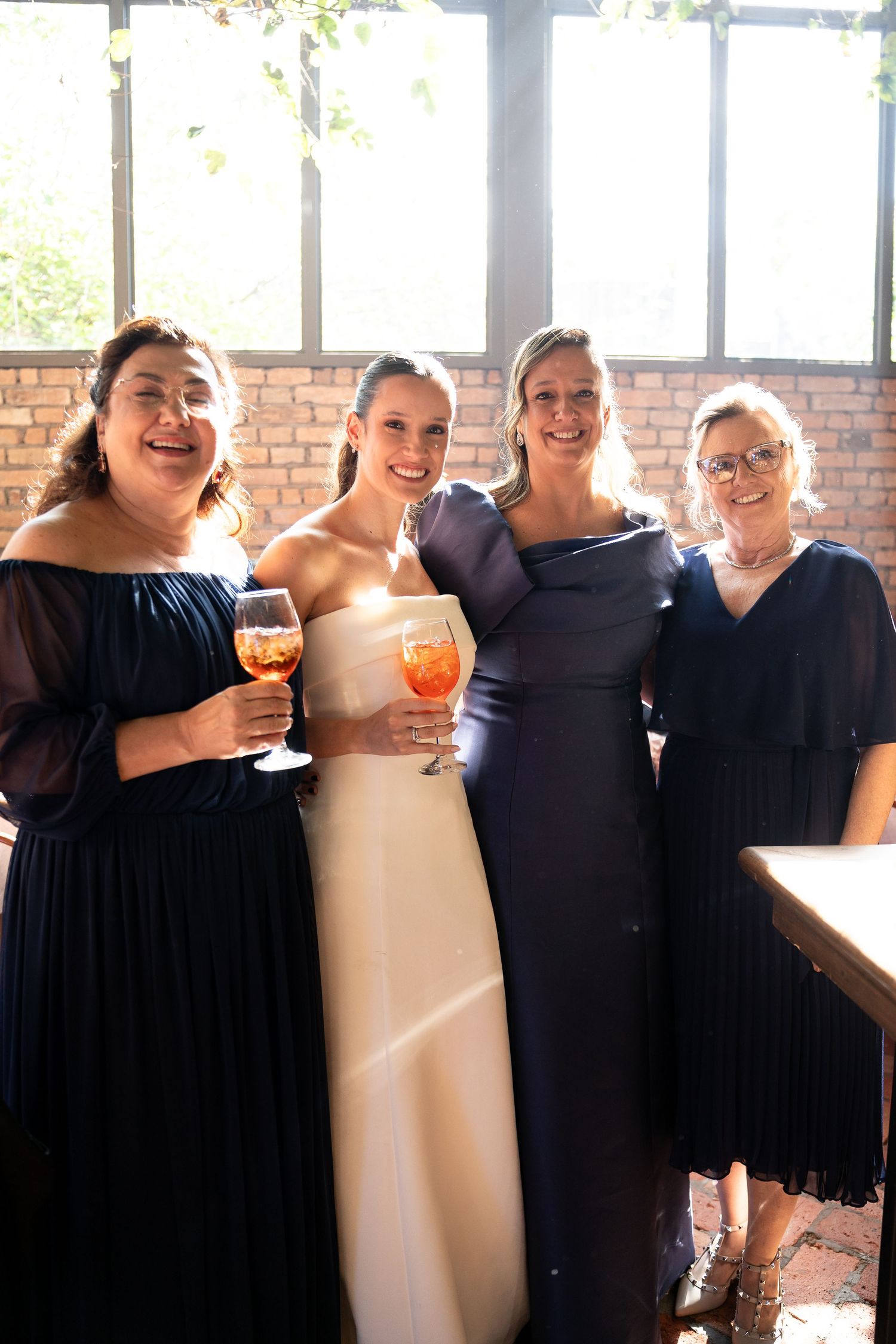 Bride in white gown poses with three bridesmaids in navy blue dresses, holding Aperol Spritz cocktails indoors.