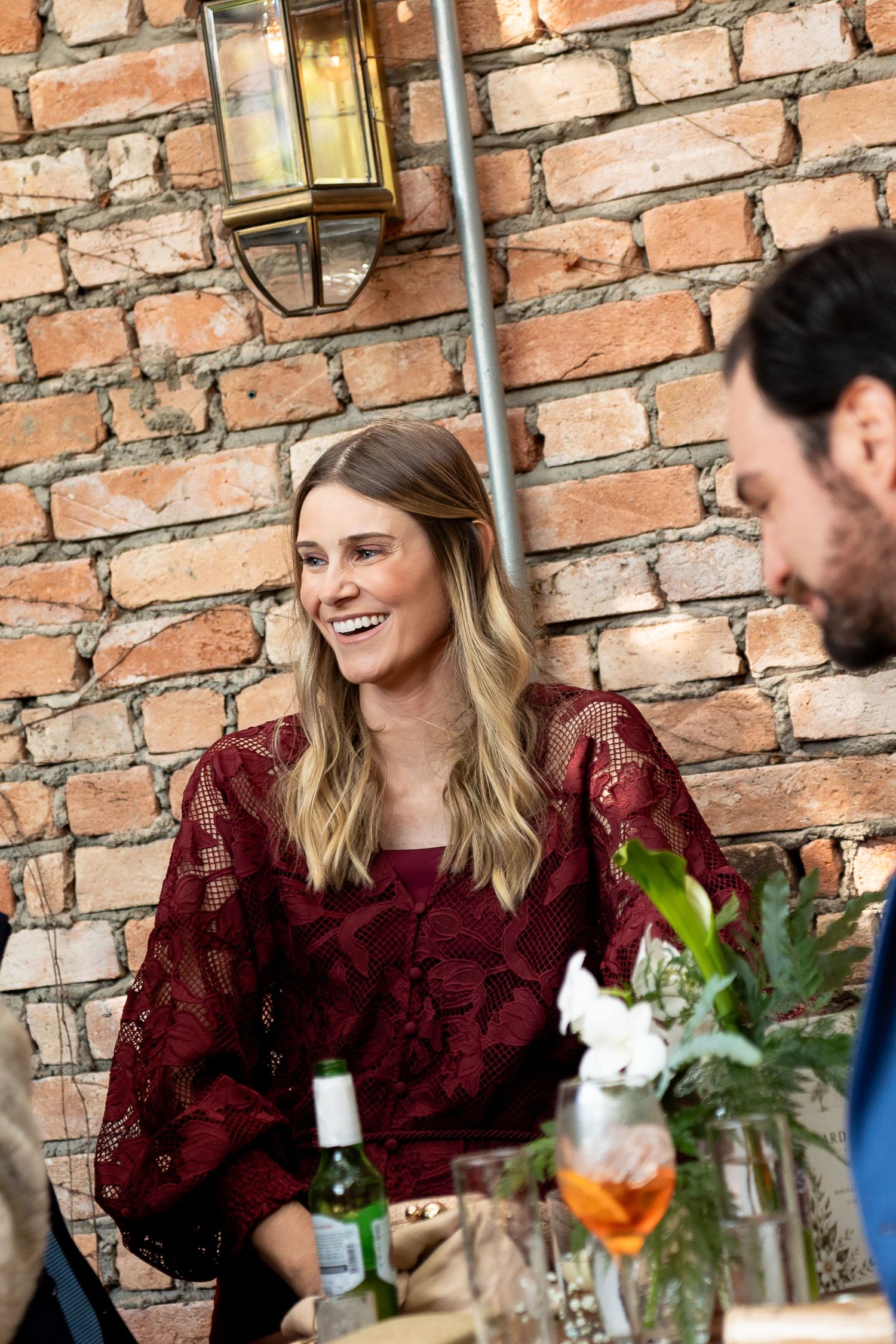 Woman in burgundy lace dress smiling at outdoor dining table against brick wall.