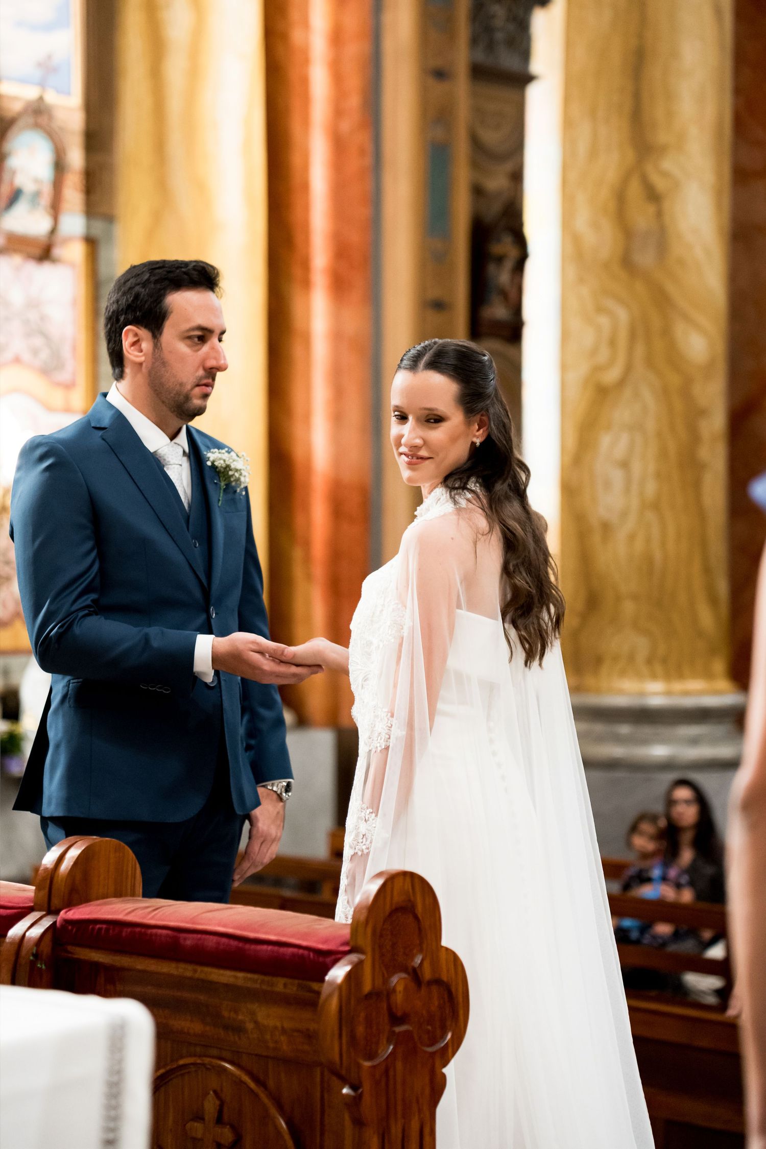Bride and groom exchange vows at church altar, bride in white gown with cape, groom in navy suit.