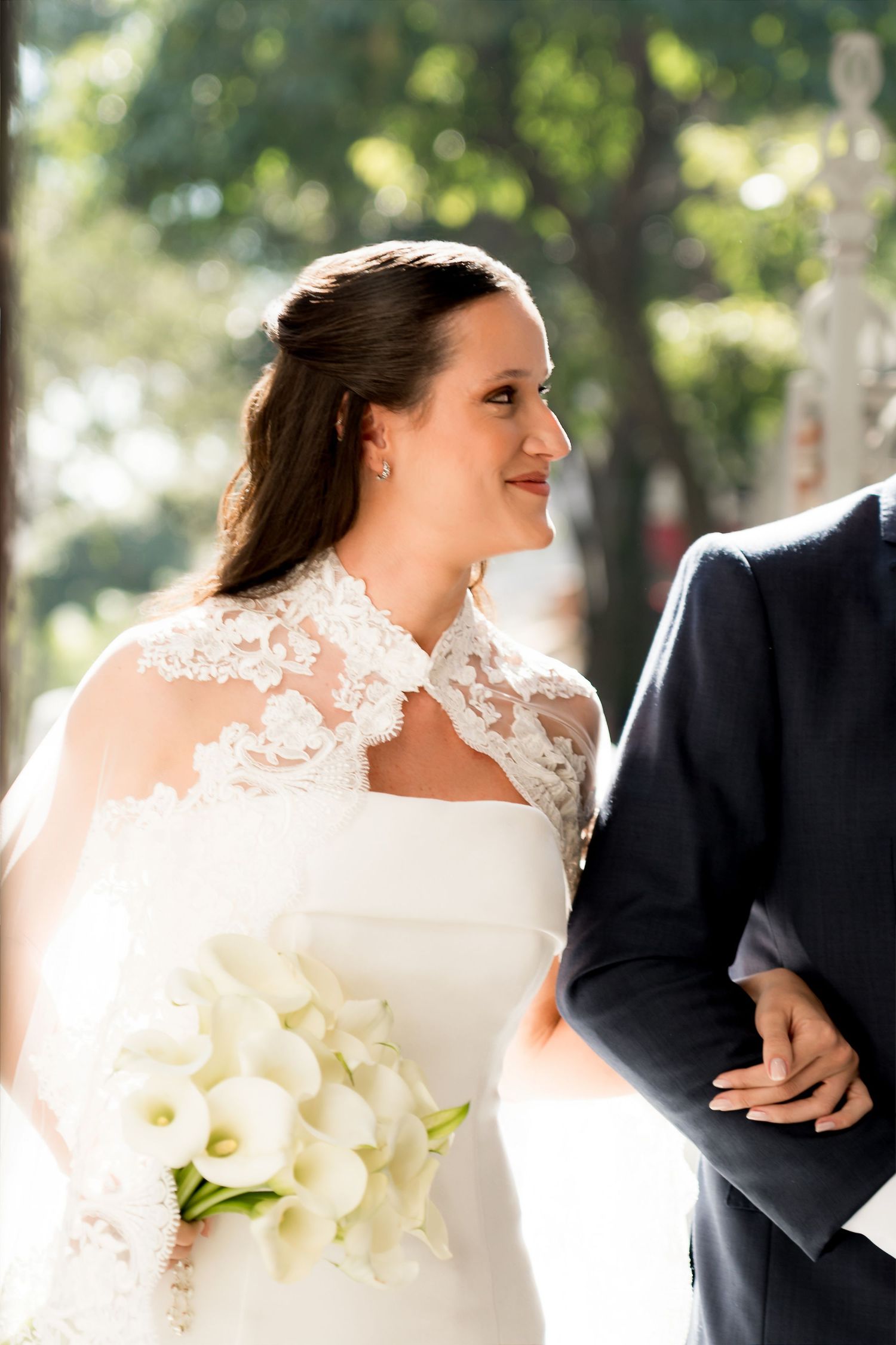 Bride in lace wedding dress holding white calla lily bouquet smiles at groom in black suit outdoors.