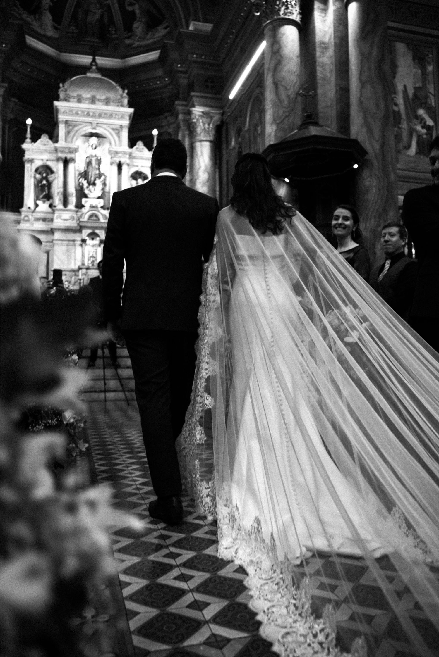 Black and white photo of a bride and groom walking down the church aisle, her lace veil trailing beautifully behind.
