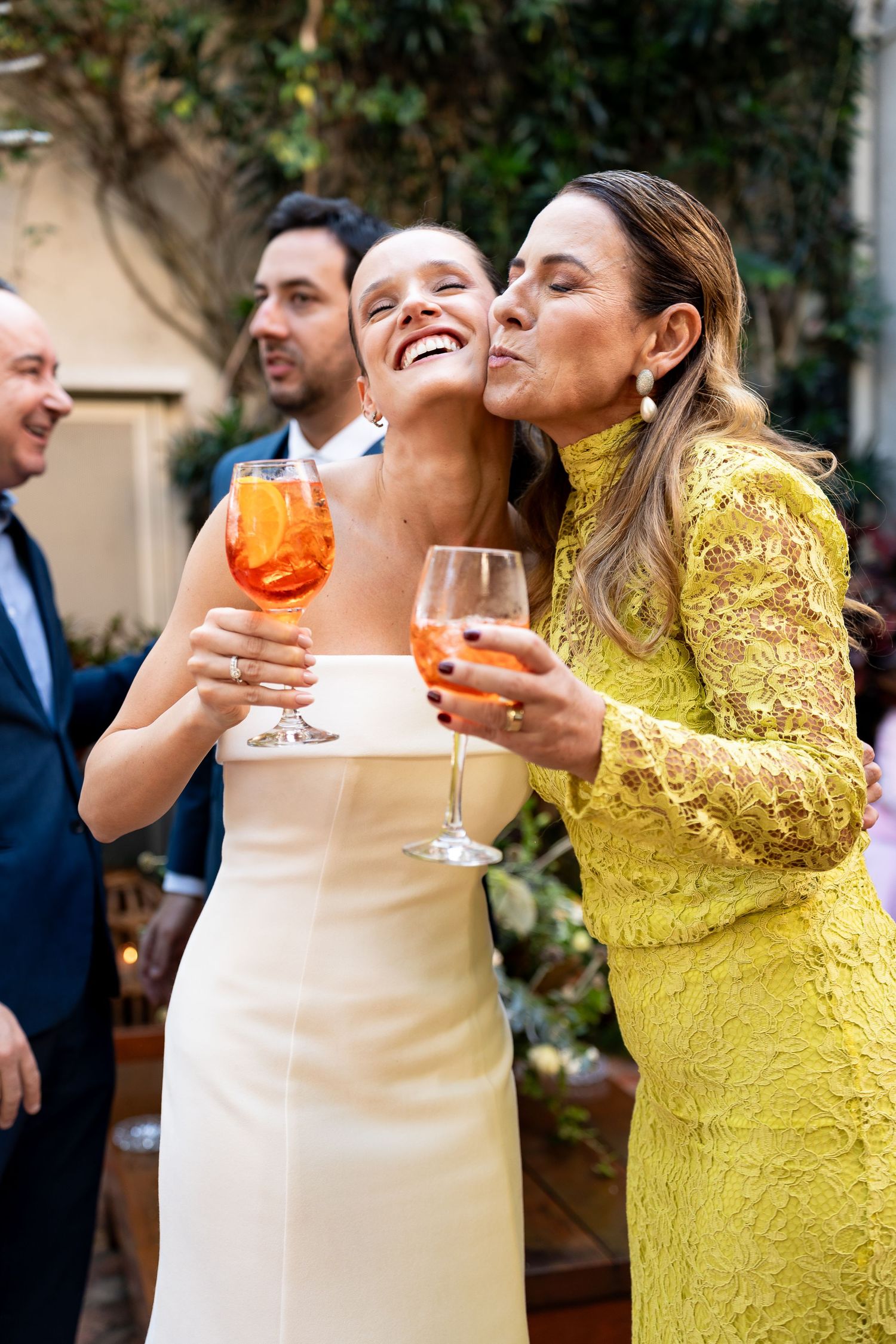 Two women in elegant attire share a joyful moment, holding wine glasses at a wedding celebration outdoors.
