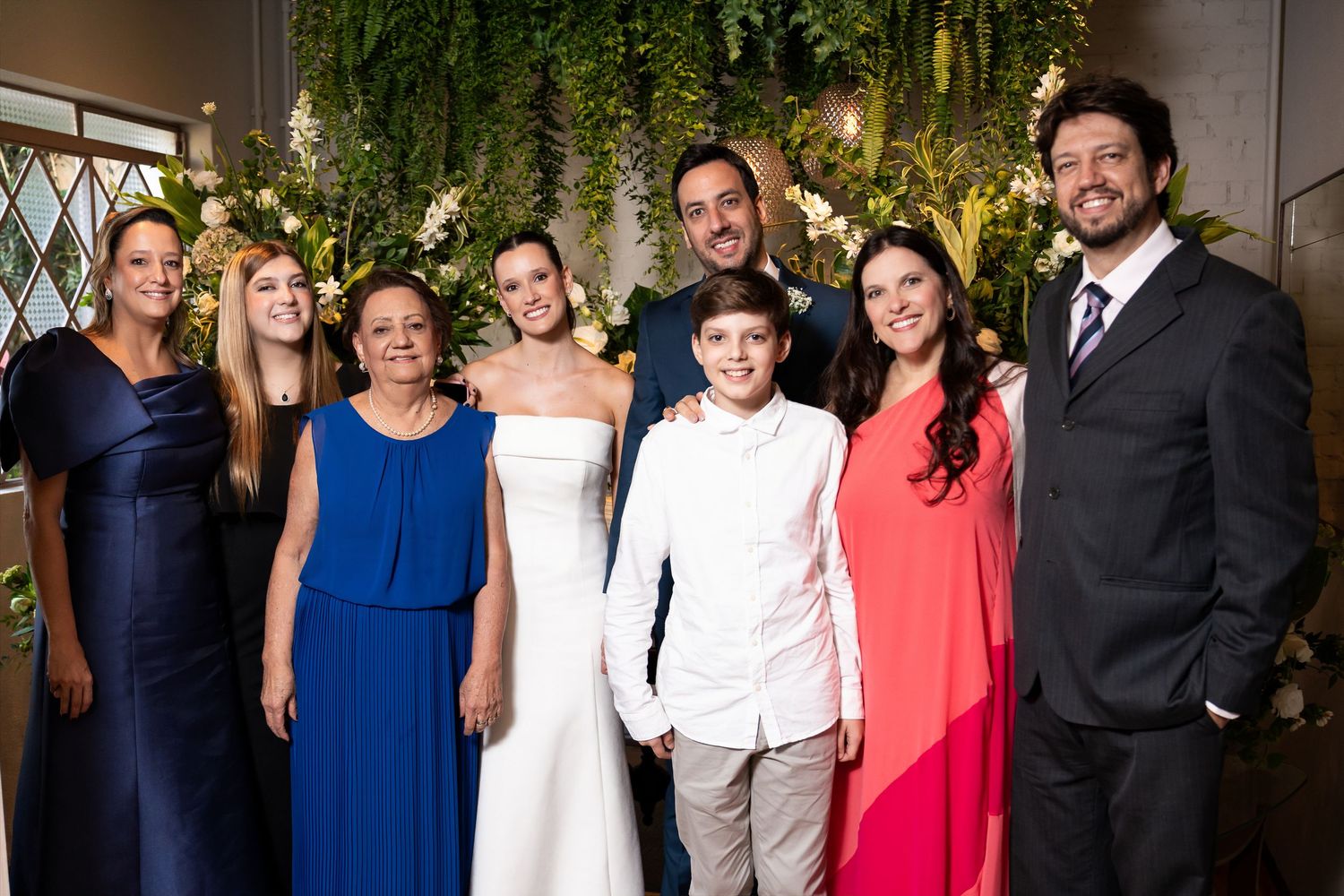 Wedding party group photo with bride in white gown, groom, and family members posing before floral backdrop.