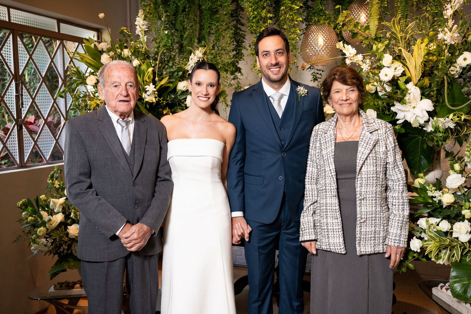 Bride and groom pose with elderly couple in front of floral backdrop at elegant wedding ceremony.