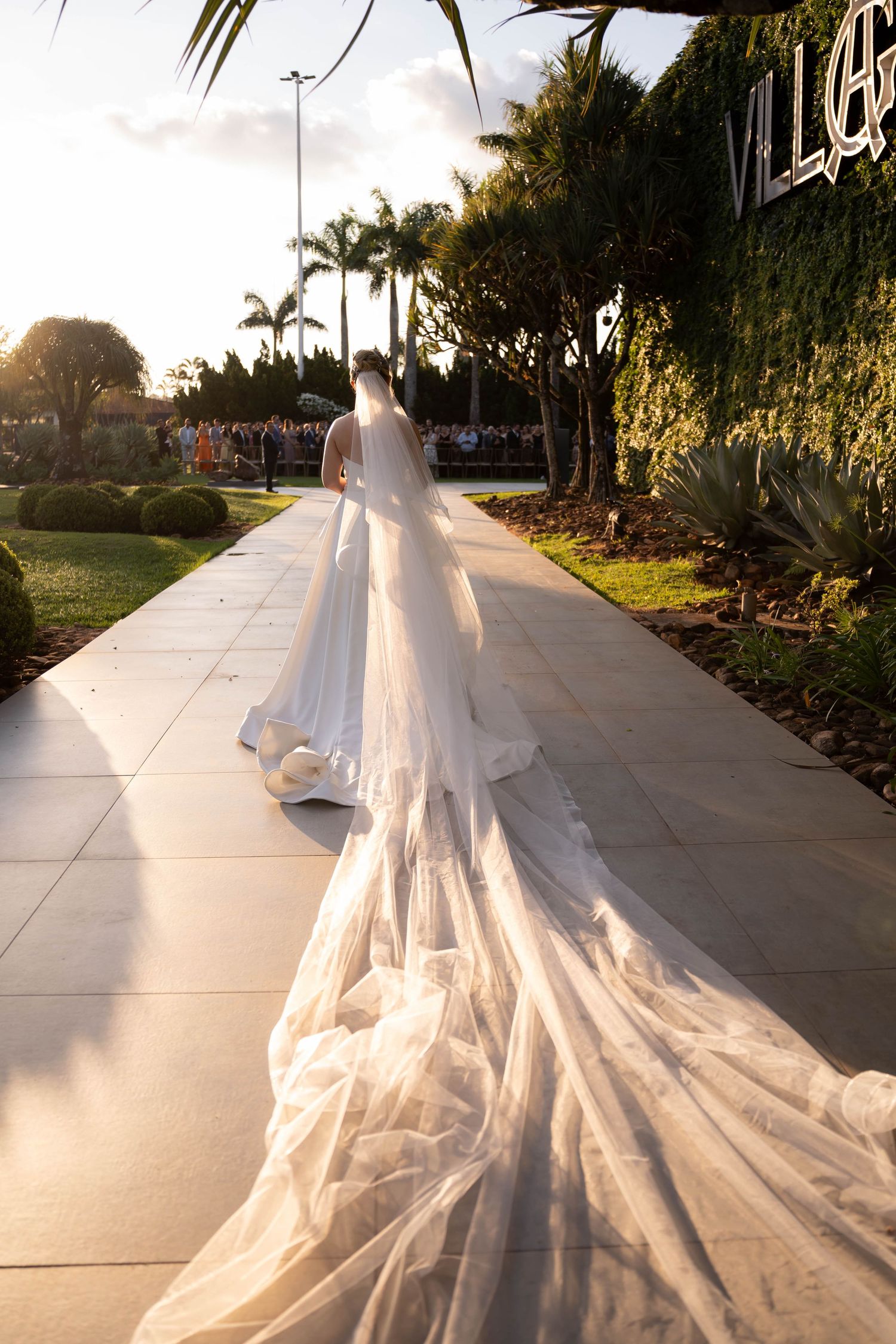 Bride walks down outdoor path in stunning white wedding gown with an extraordinarily long flowing veil trailing behind her.