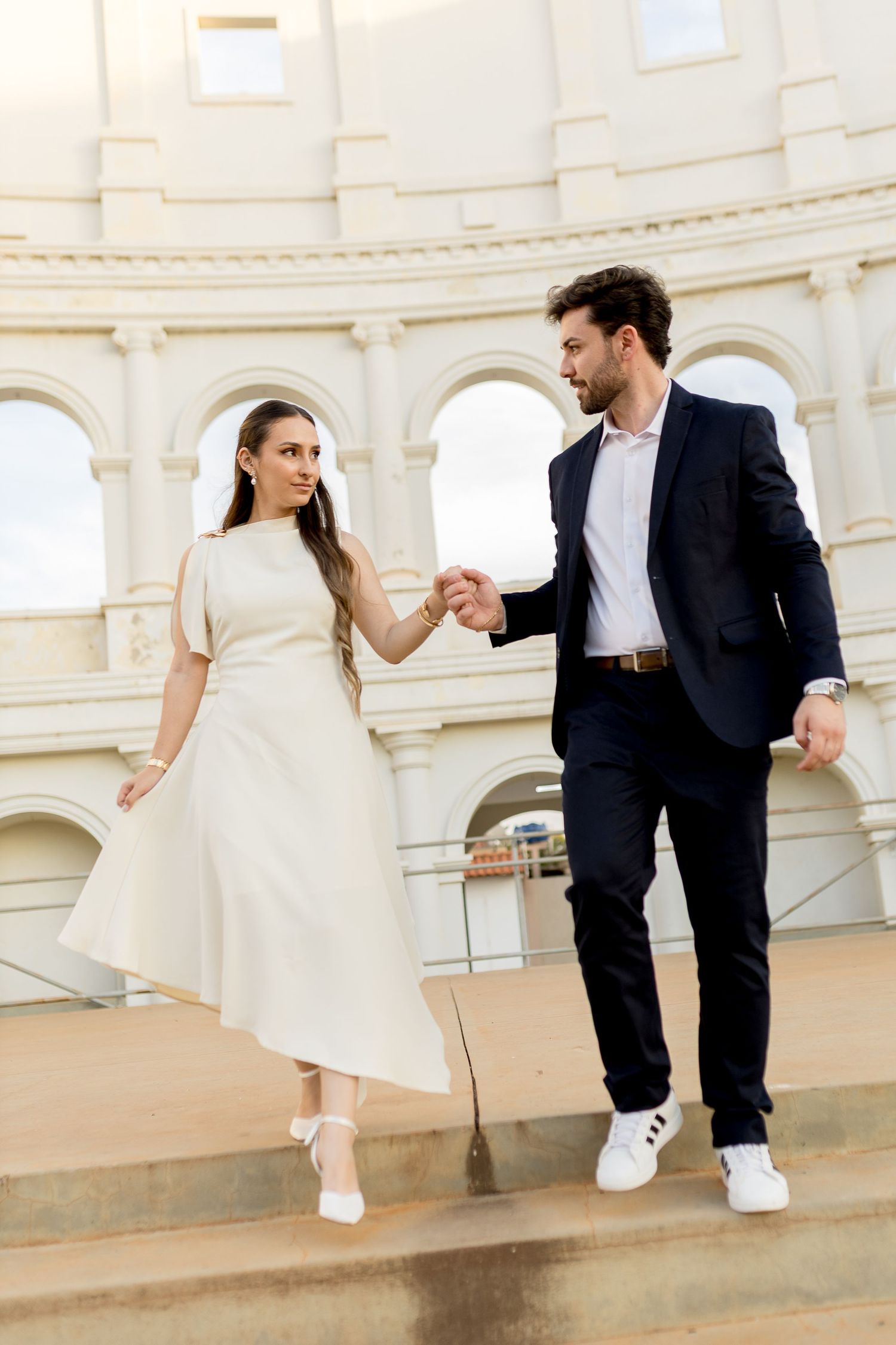 Elegant couple holding hands on steps before classical arched building, woman in white dress, man in navy suit.