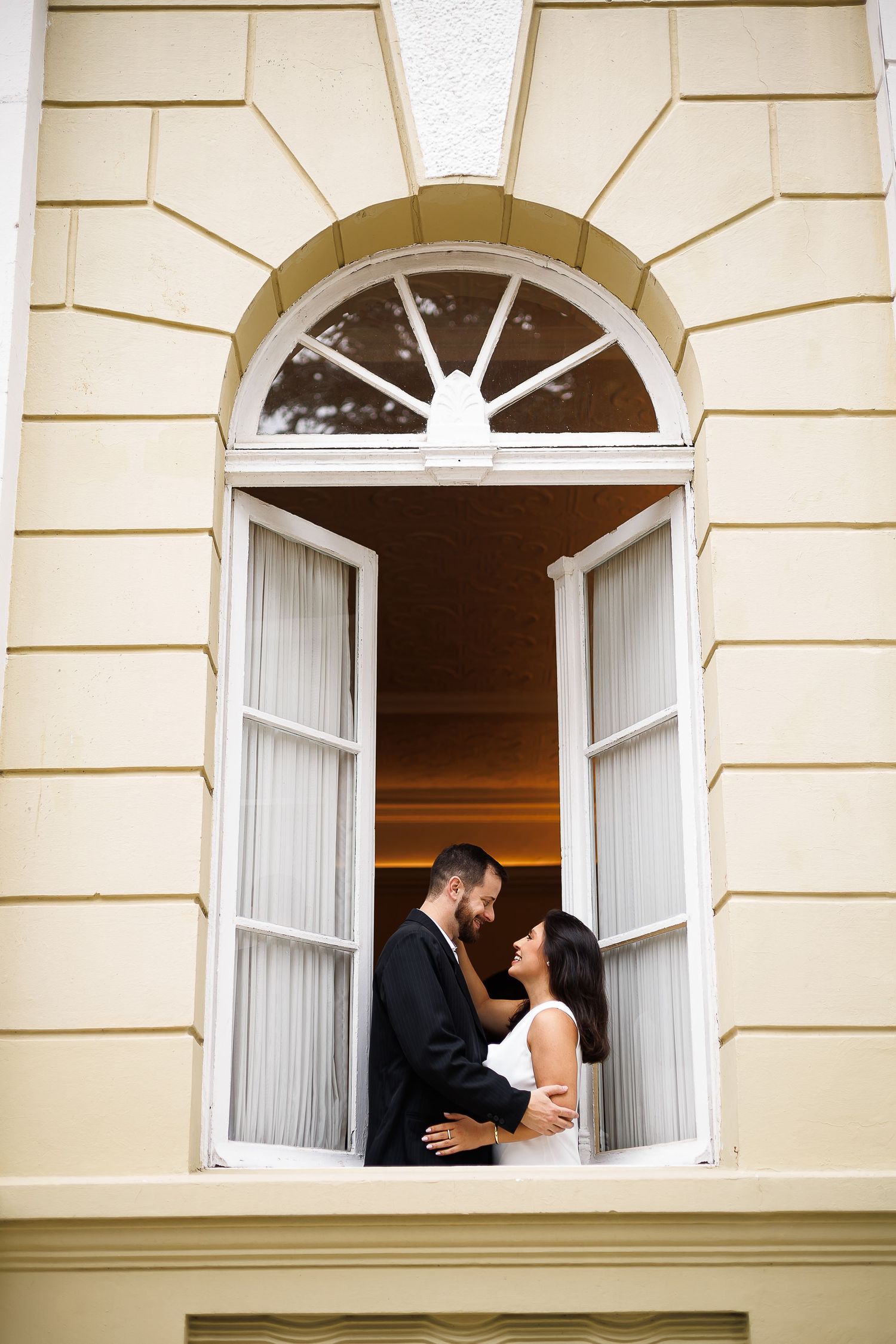 Bride and groom share a romantic moment framed in an elegant arched window of a classical building.