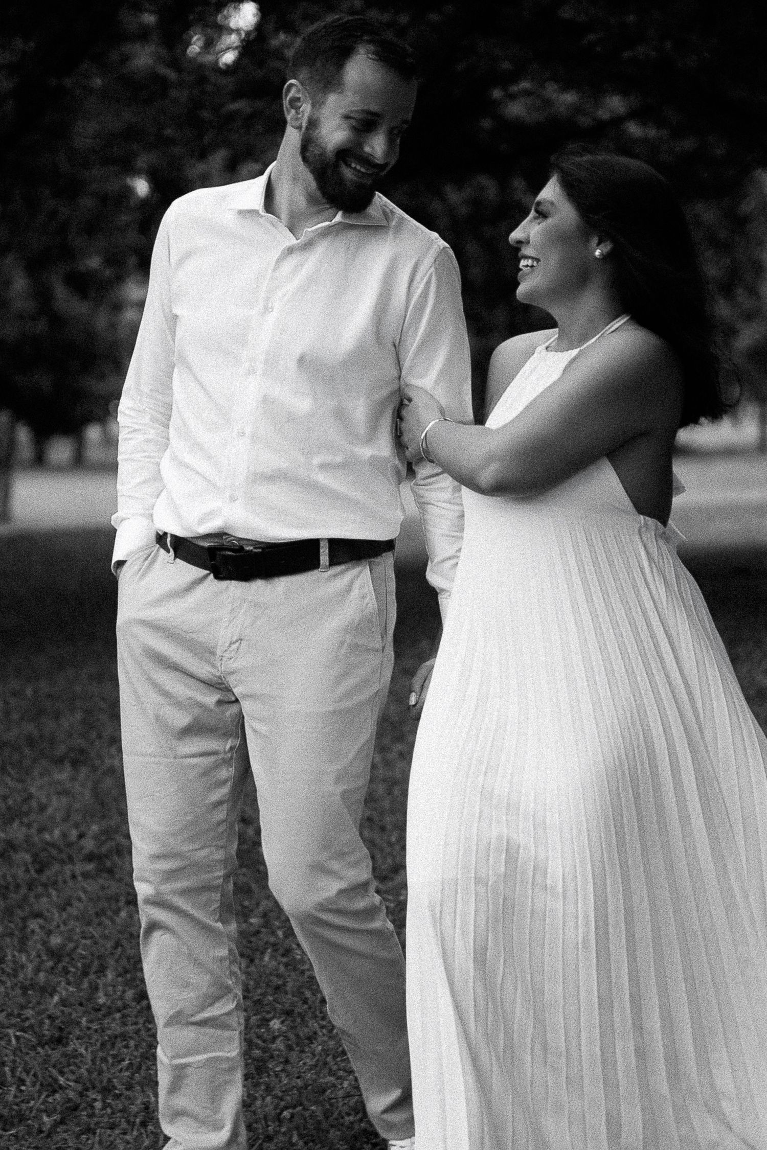 Black and white photo of a smiling couple in white outfits walking together in a park, sharing a joyful moment.