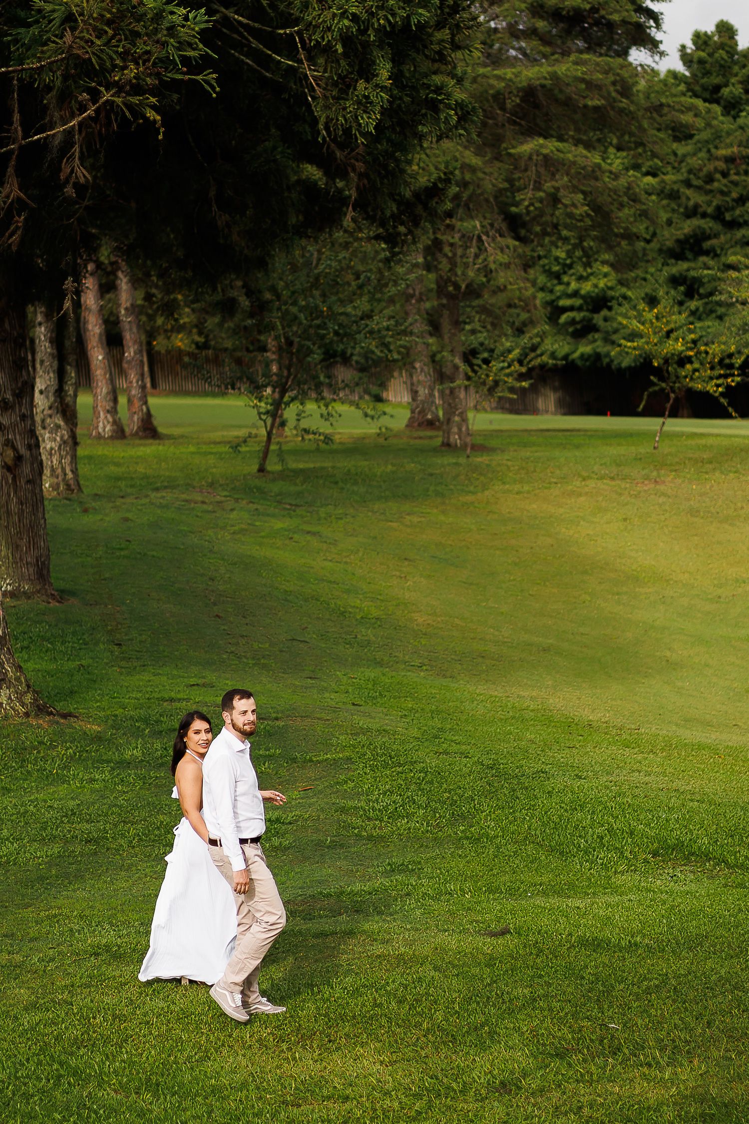 Bride and groom walking hand in hand across lush green lawn surrounded by tall trees during outdoor wedding.