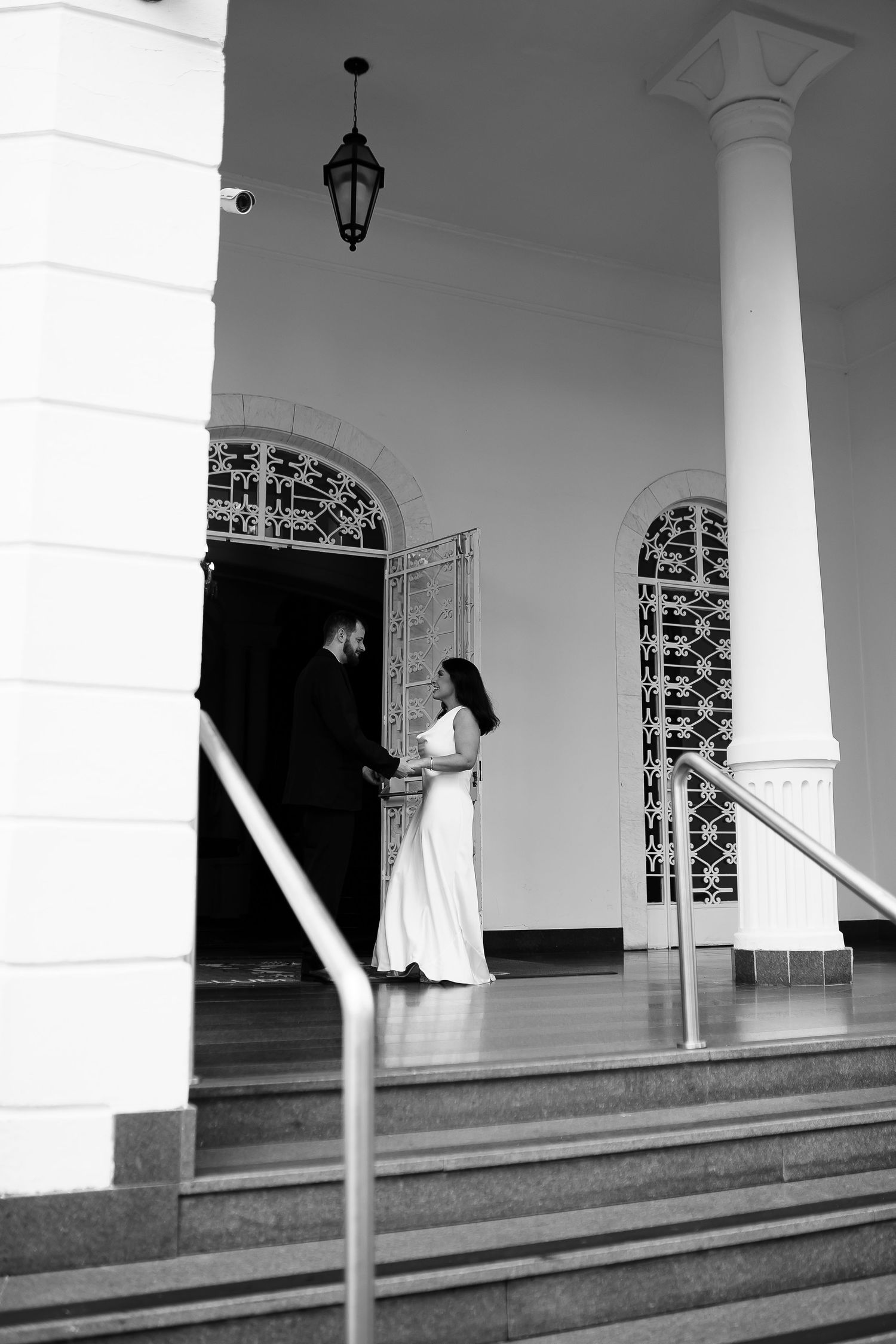 Black and white photo of a bride and groom standing at the entrance of an elegant white building with columns.