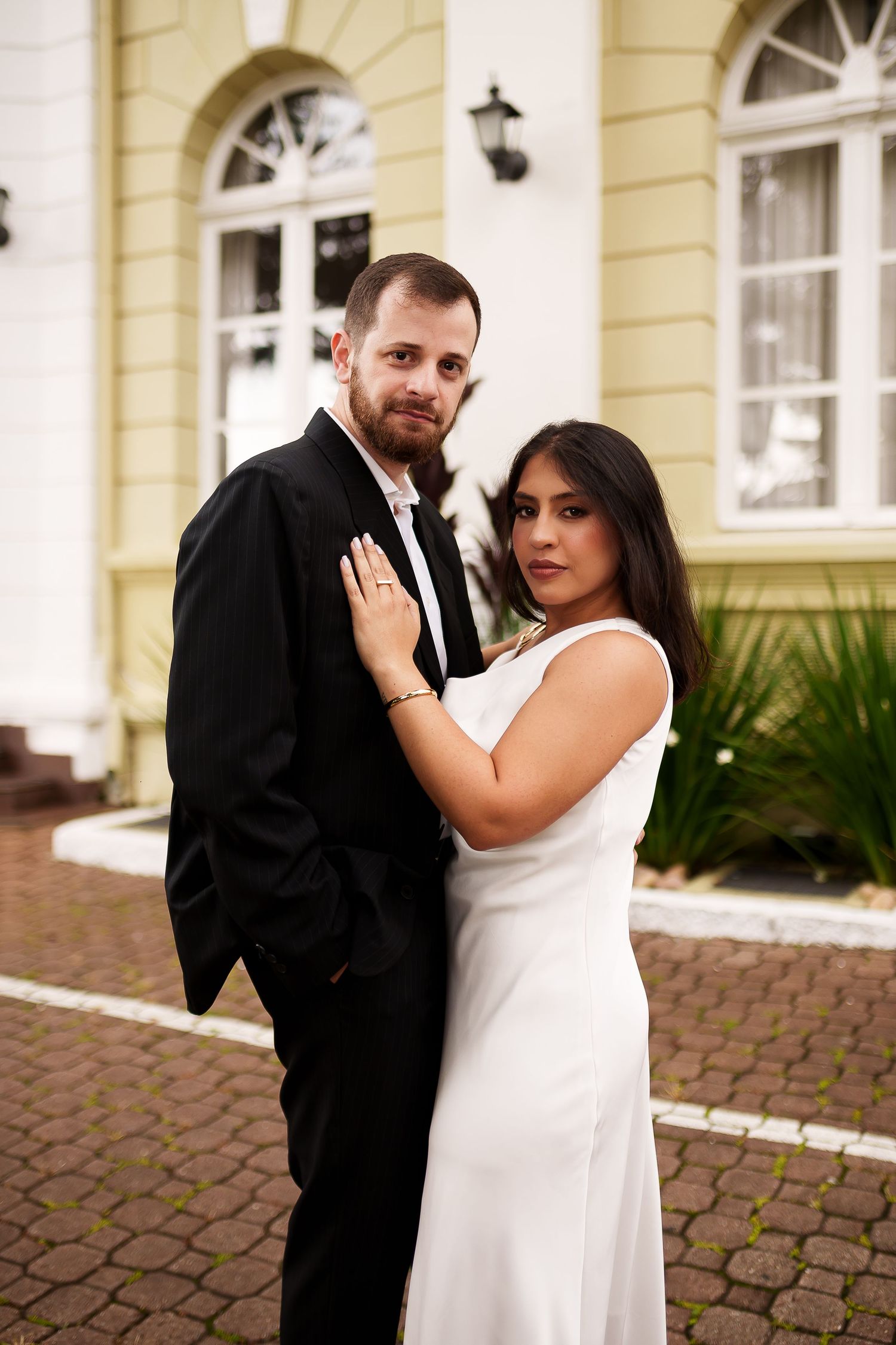Bride in white dress and groom in black suit posing together outside an elegant yellow building.