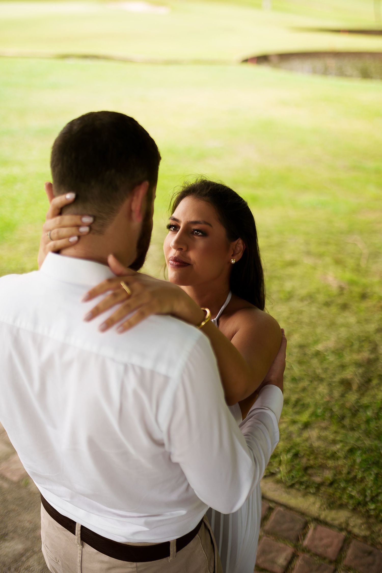 A couple embraces outdoors, woman in white dress gazes lovingly at man in white shirt on green lawn.