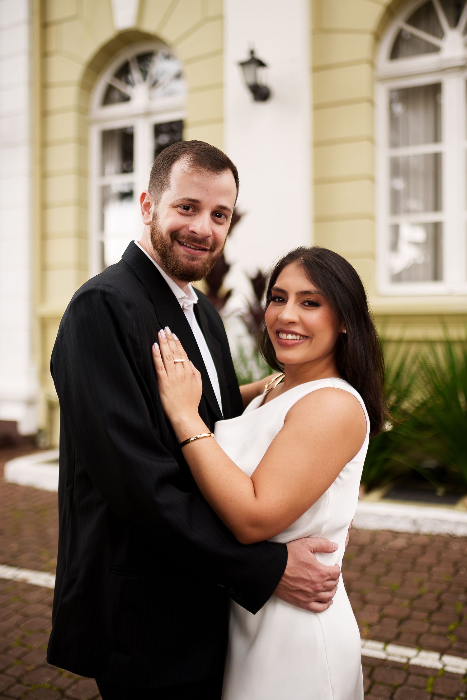 Smiling couple in wedding attire embracing outdoors, groom in black suit and bride in white dress.