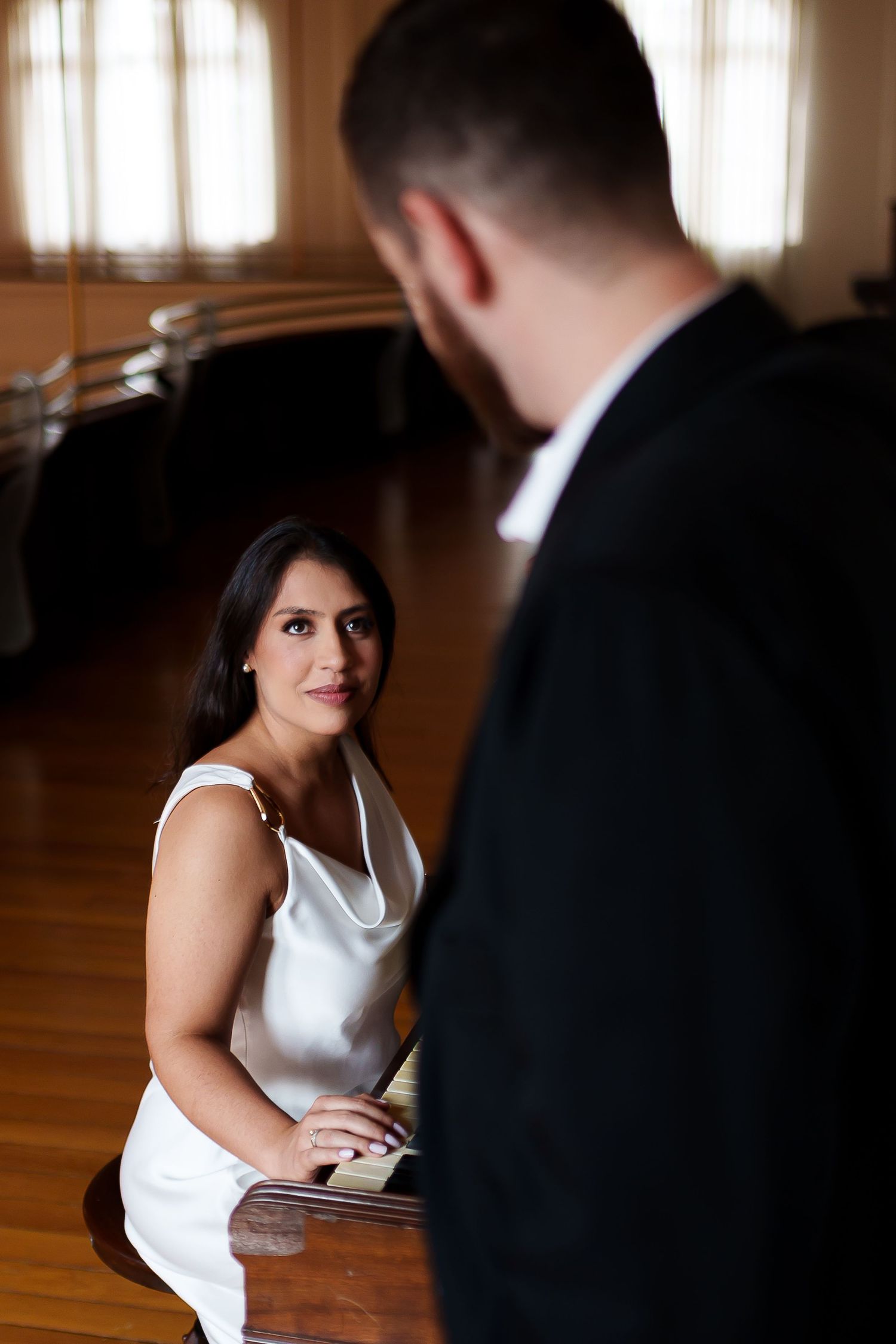 Bride in white dress sits at piano gazing at groom in black suit in elegant wood-paneled room.