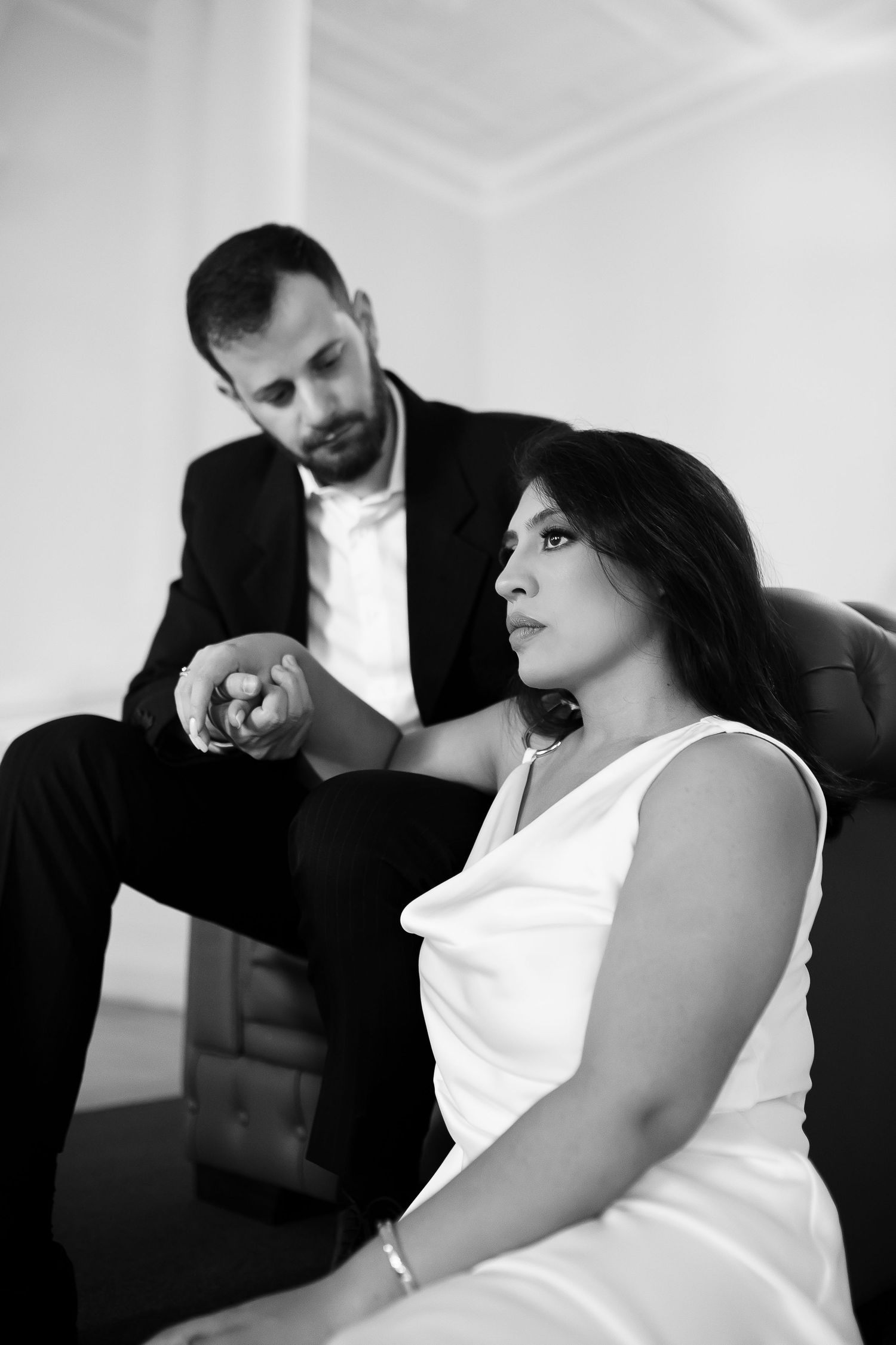 Black and white photo of a woman in white dress sitting while a suited man checks his watch behind her.