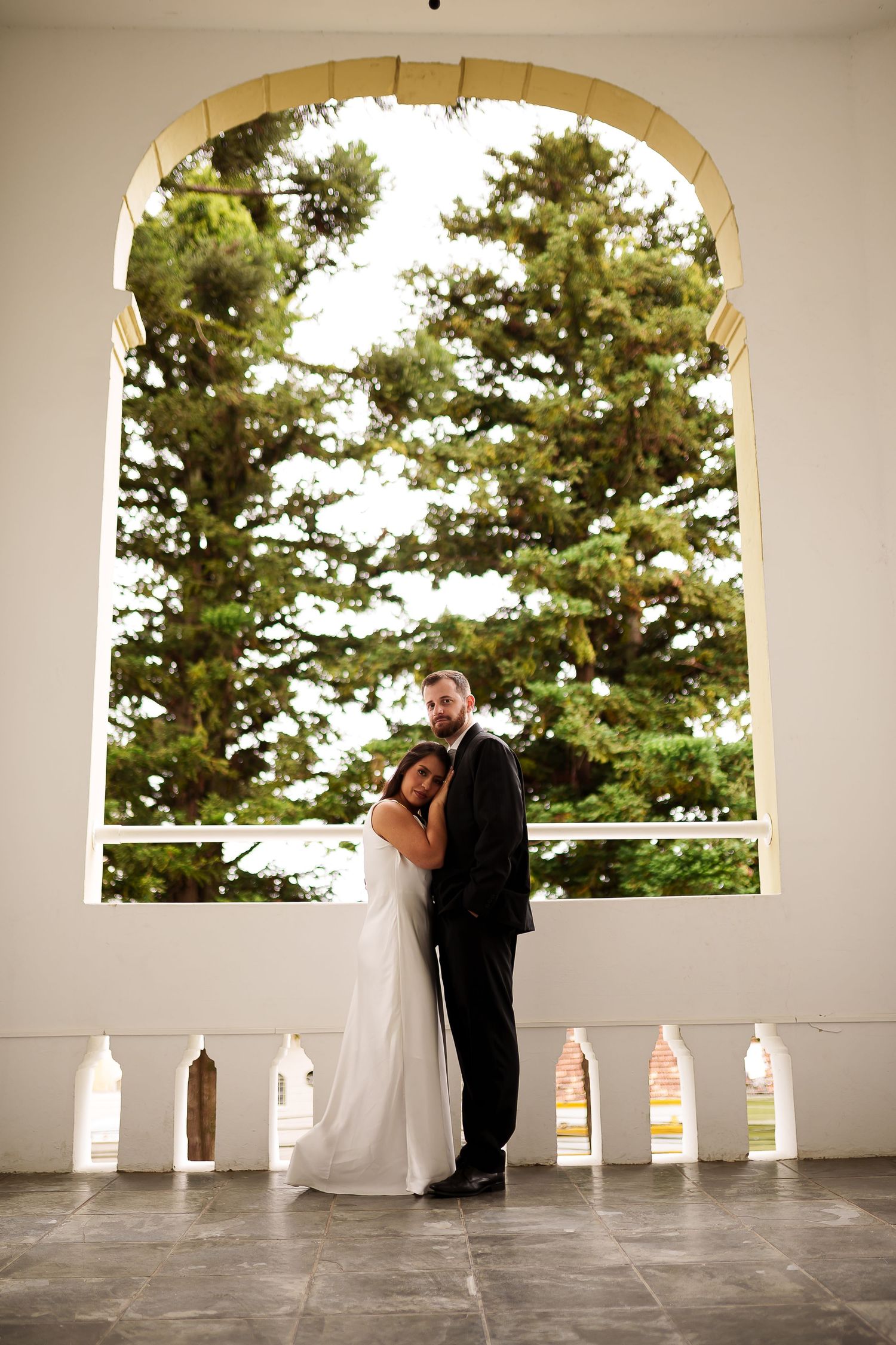 Bride and groom embracing under a grand arched window with lush green trees in the background at their wedding.