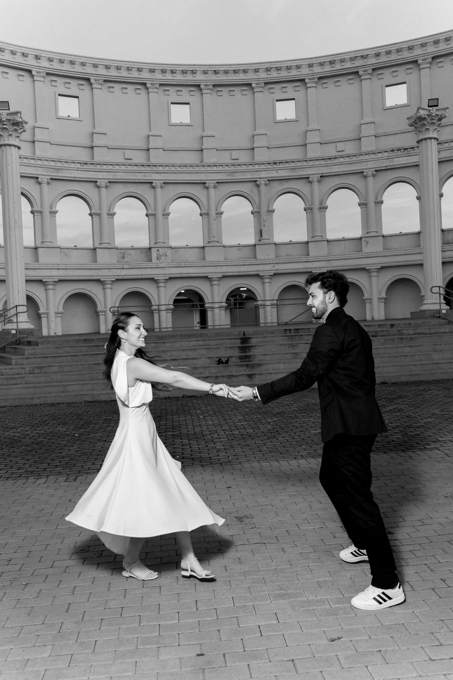 Black and white photo of a couple dancing and holding hands in front of a Roman Colosseum replica.