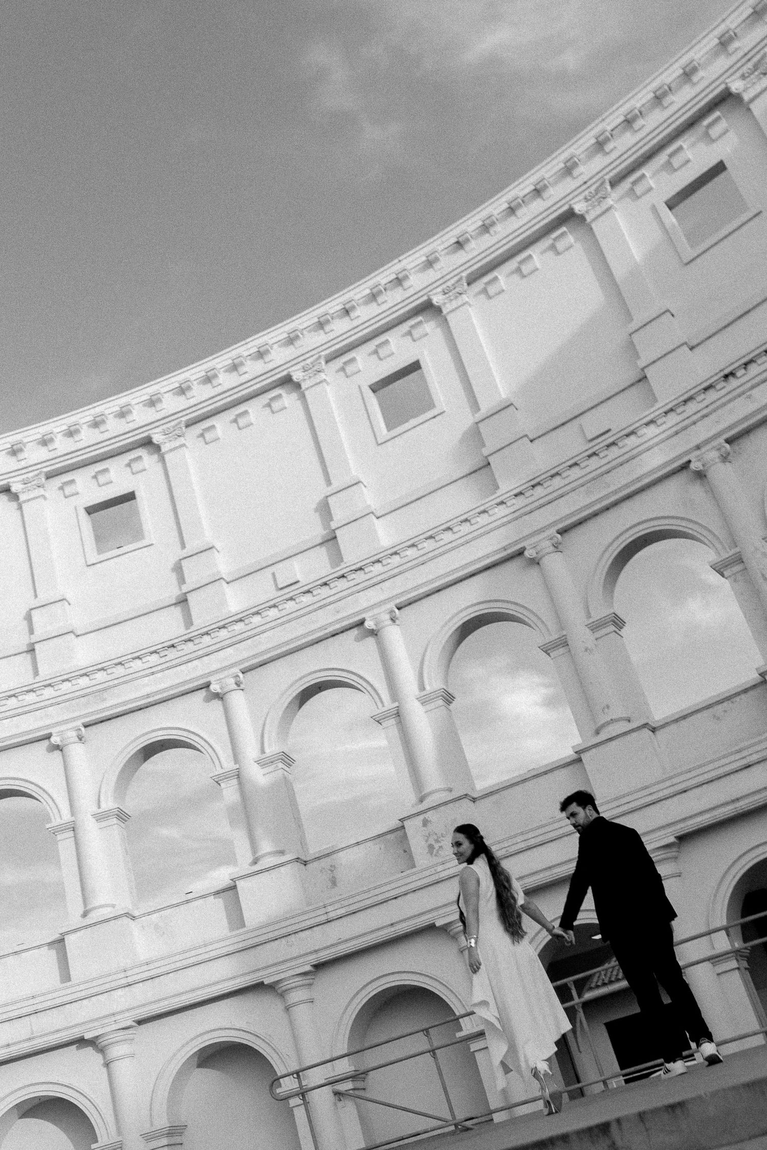 Black and white photo of a couple in wedding attire holding hands at the Roman Colosseum in Rome, Italy.