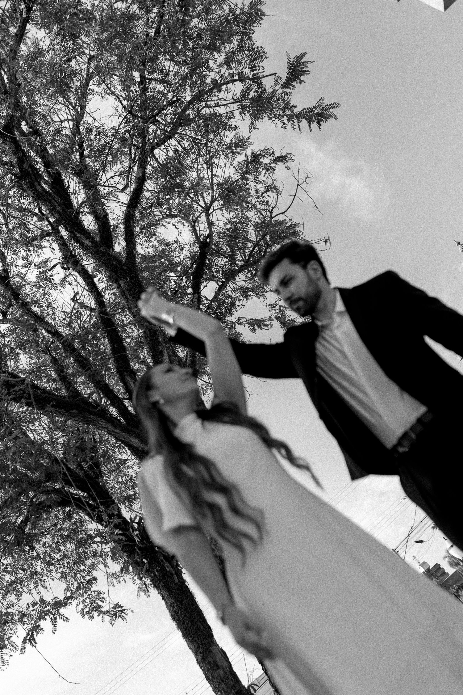 Black and white photo of a bride and groom dancing outdoors under a tree on their wedding day.
