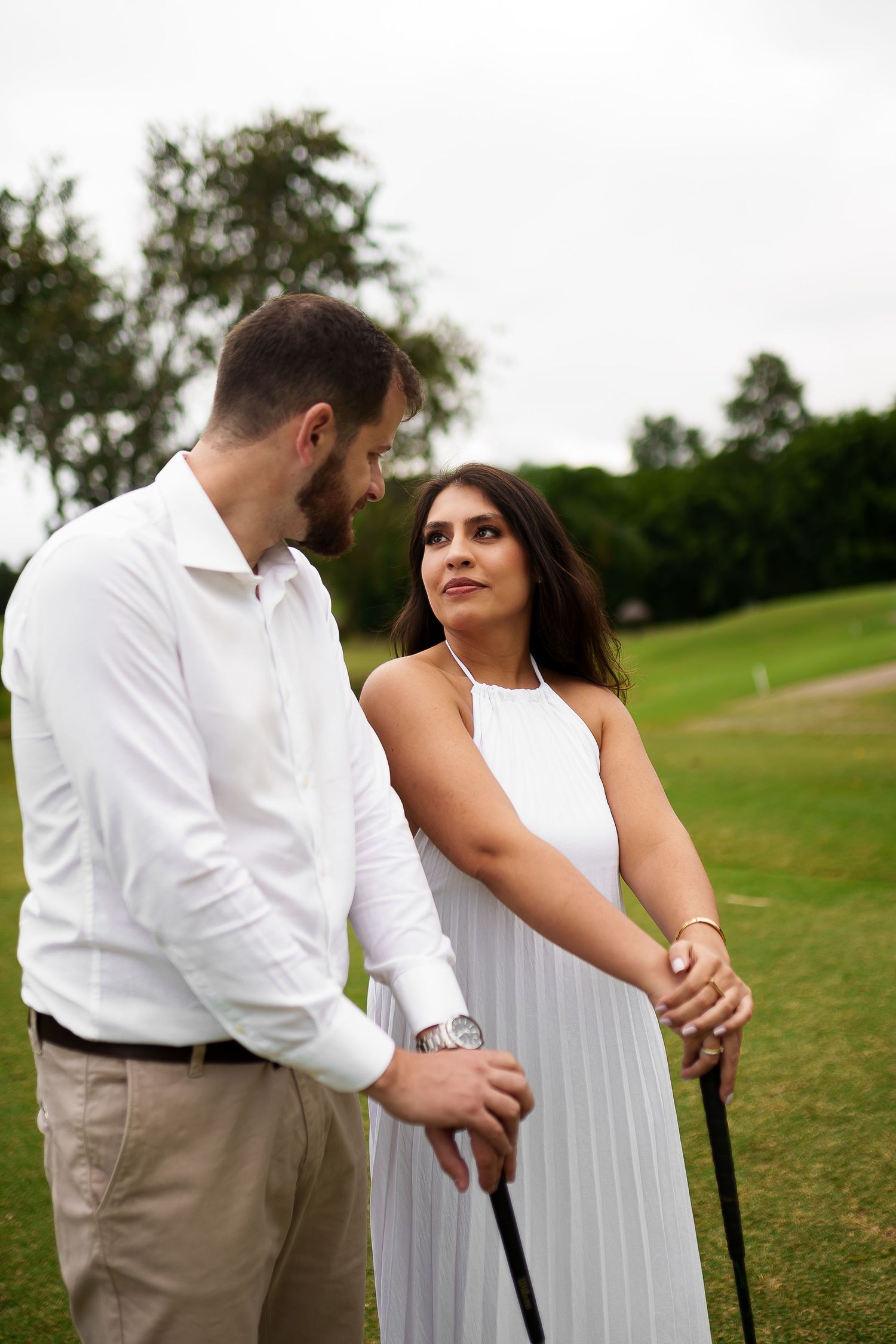 Couple in white outfits holding a golf club together on a lush green golf course, sharing a romantic moment.