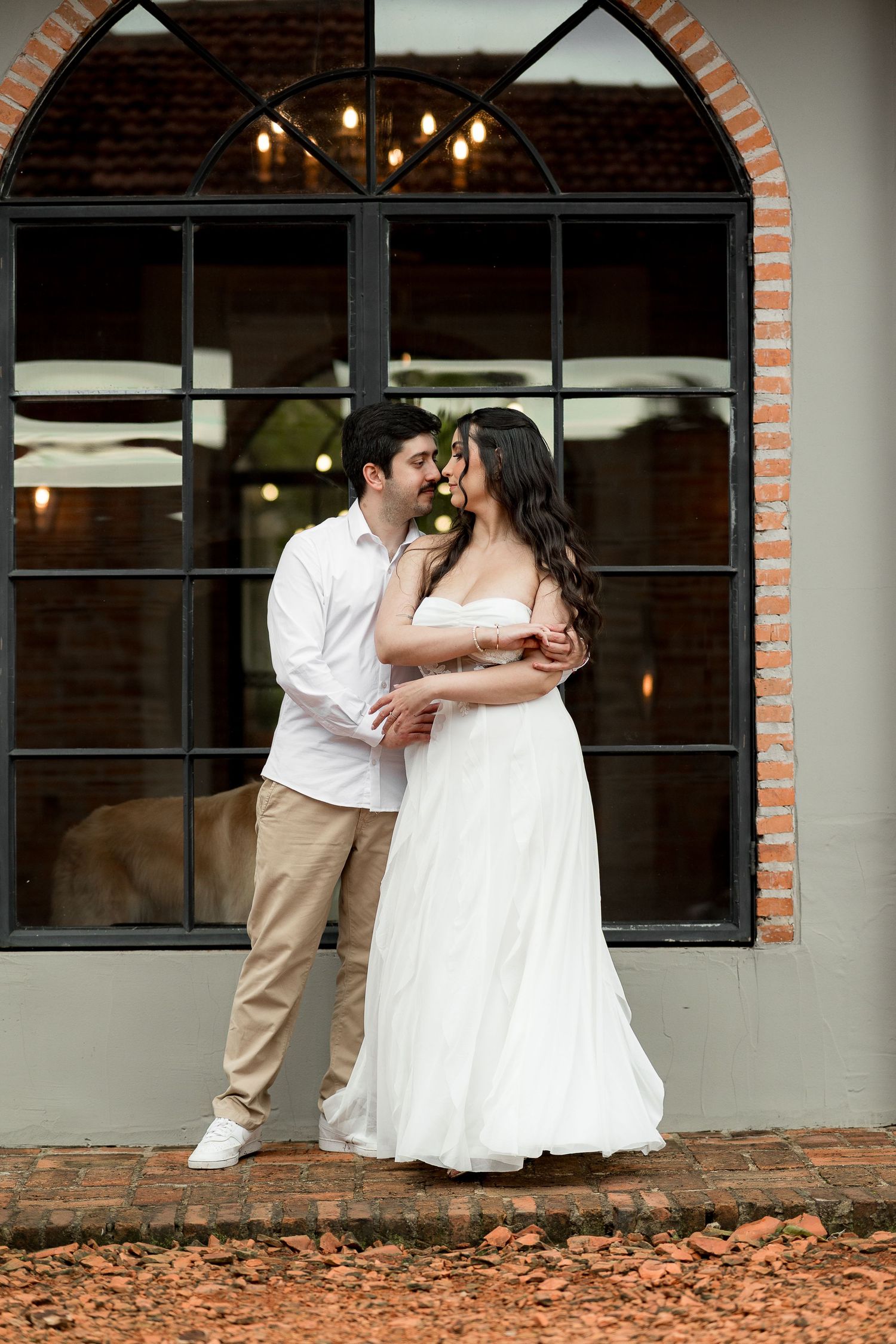 Couple in wedding attire embracing romantically in front of an arched window with warm string lights.