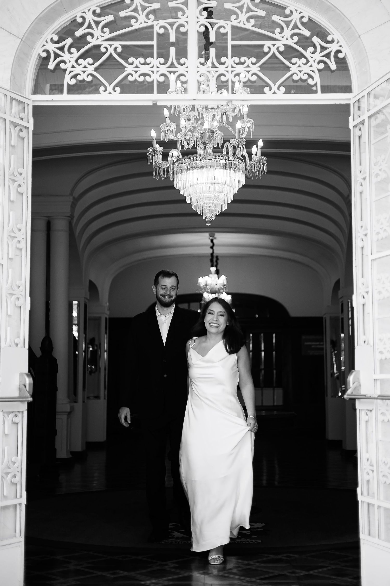 Black and white photo of a bride and groom walking through an elegant arched hallway with a crystal chandelier.
