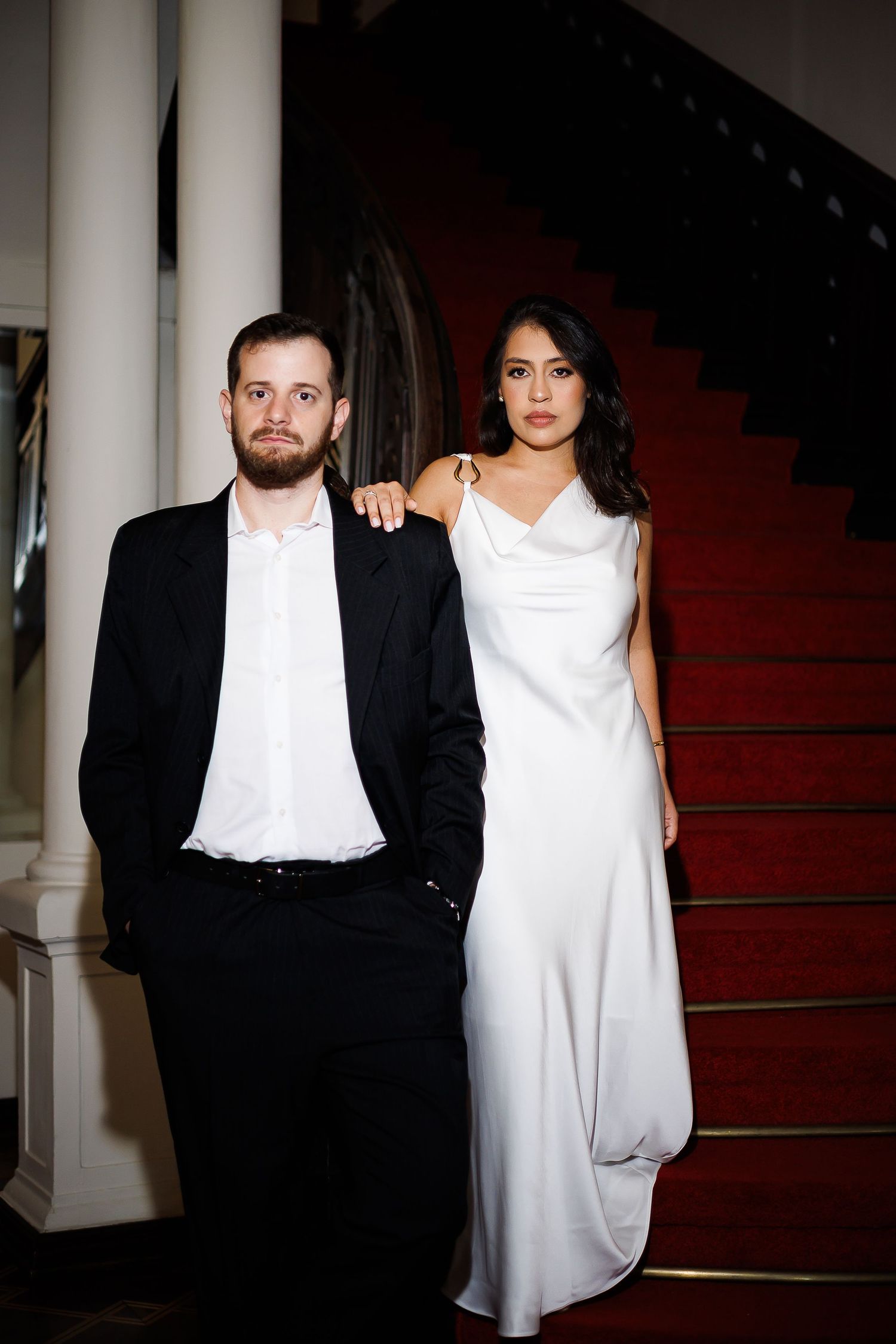 Elegant couple posing on red carpeted staircase, man in black suit, woman in white satin gown.