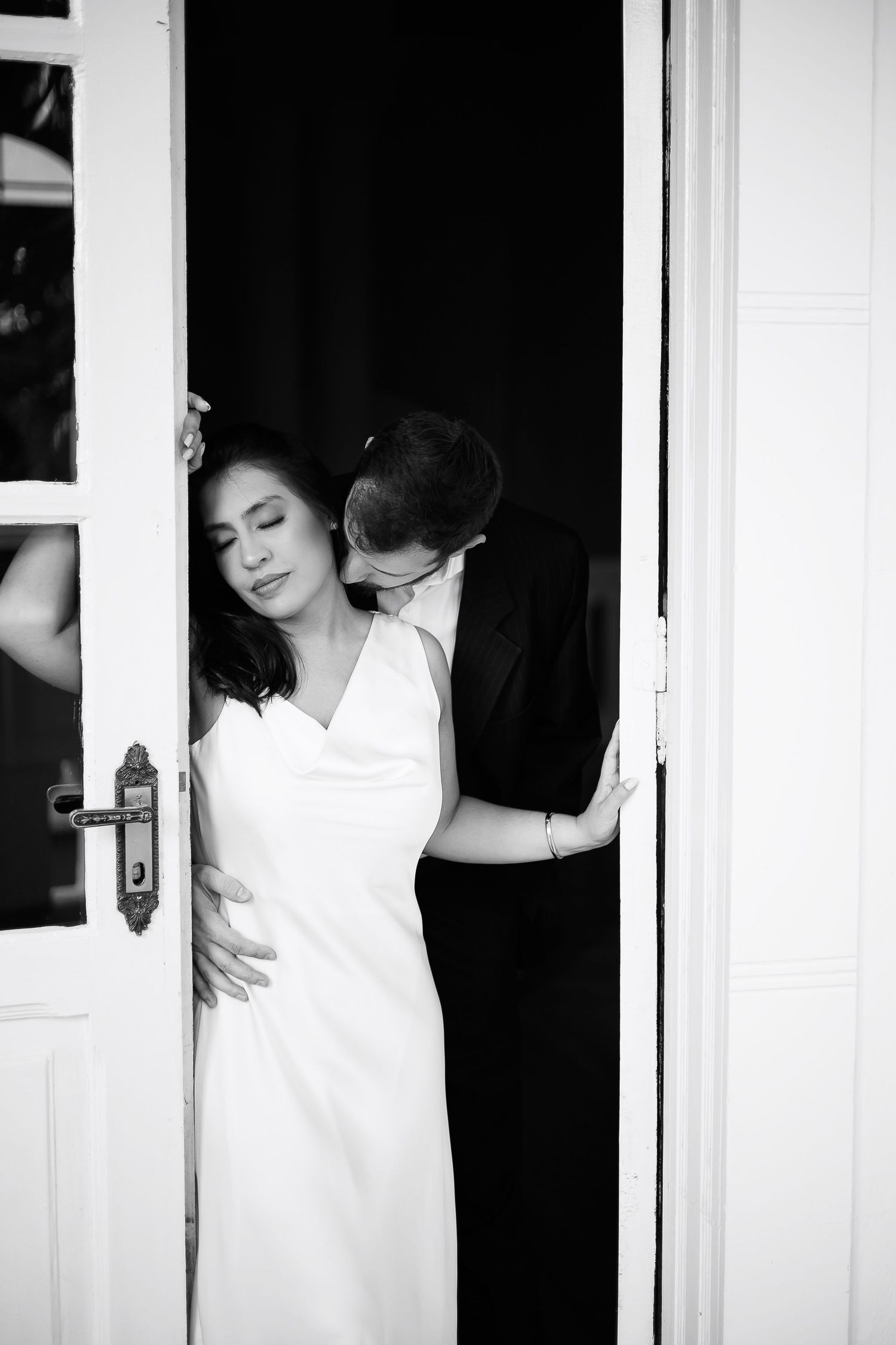 Black and white photo of a groom kissing bride on neck in doorway, bride in white dress eyes closed.