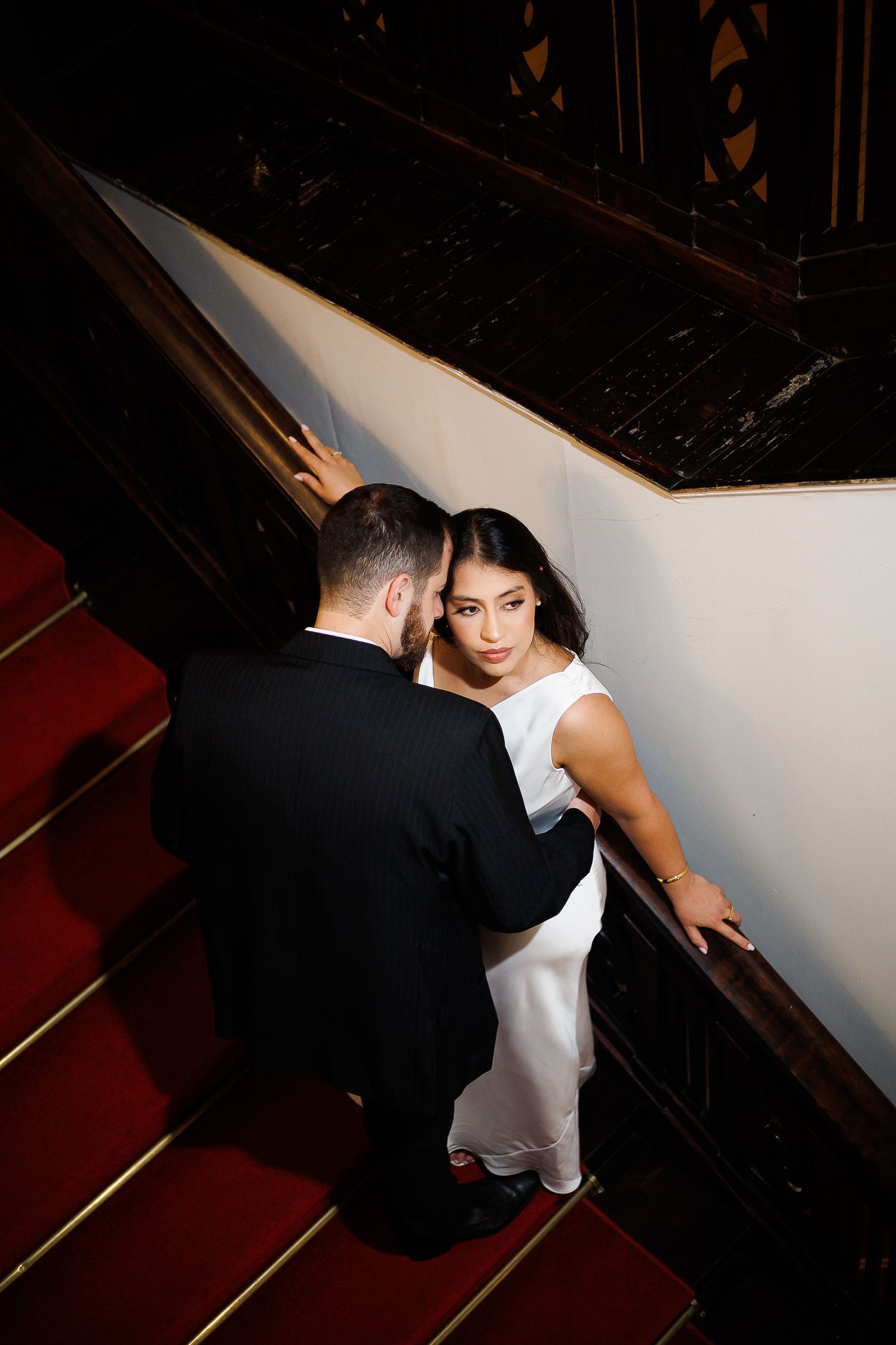 Elegant couple in formal wear on red-carpeted stairs, bride in white dress looks back while groom in black suit stands close.