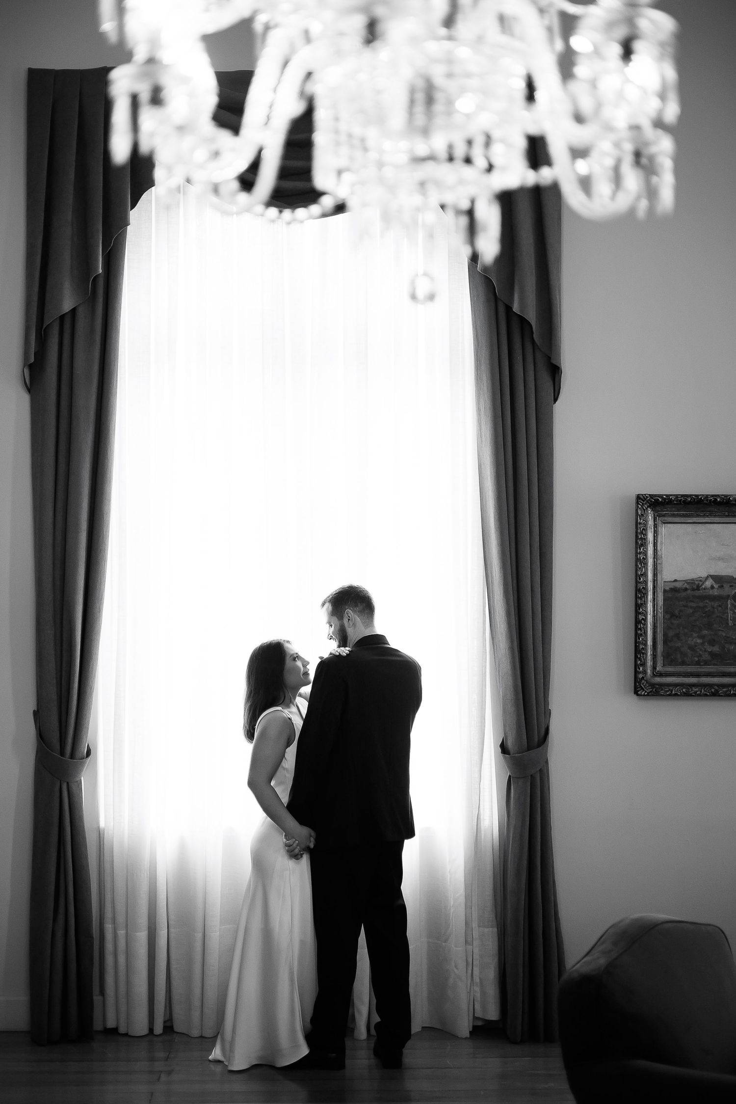 Black and white photo of an elegant couple standing by a large window beneath a crystal chandelier in a grand room.
