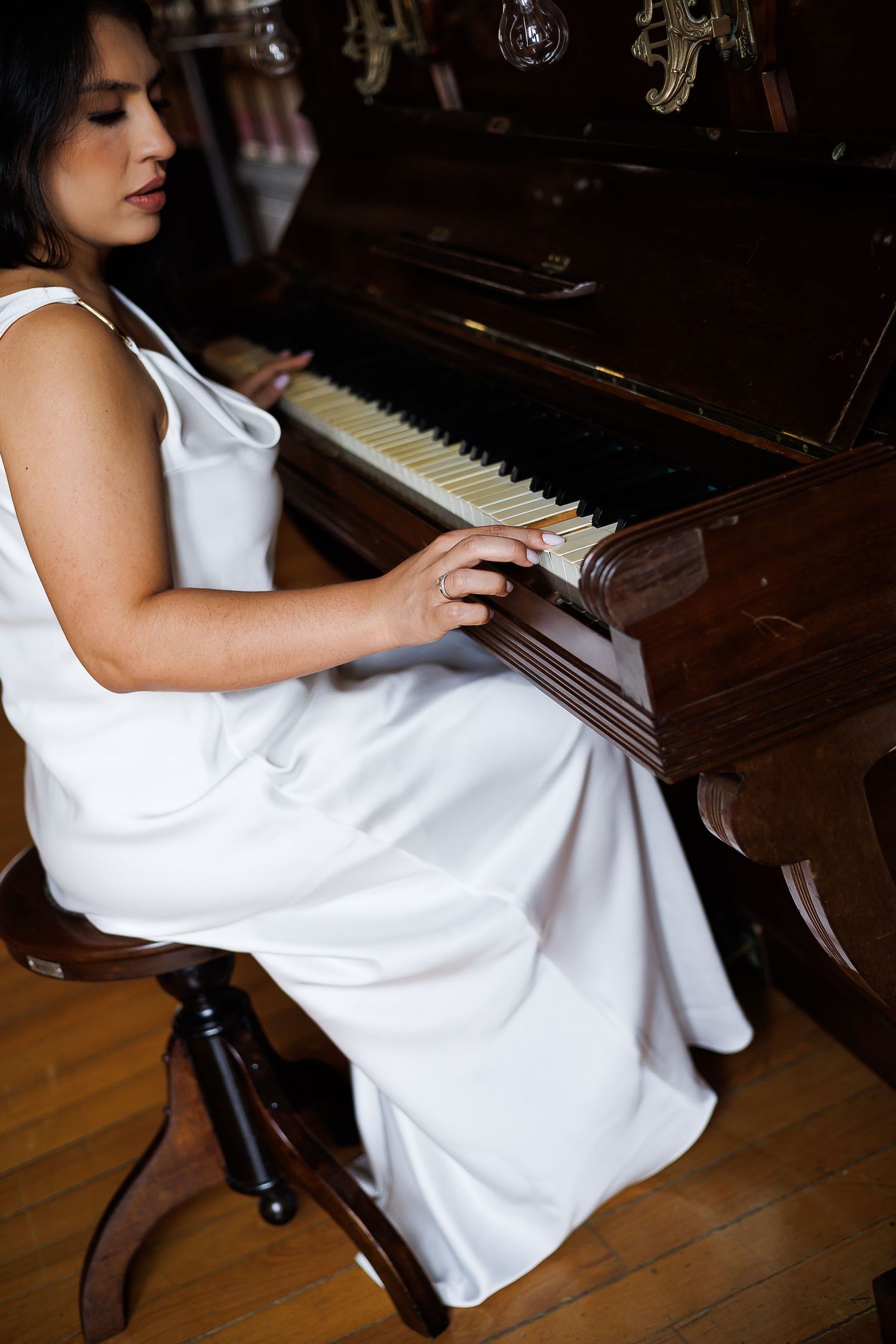 Woman in white dress playing an antique upright piano, seated on a wooden stool in a warmly lit room.