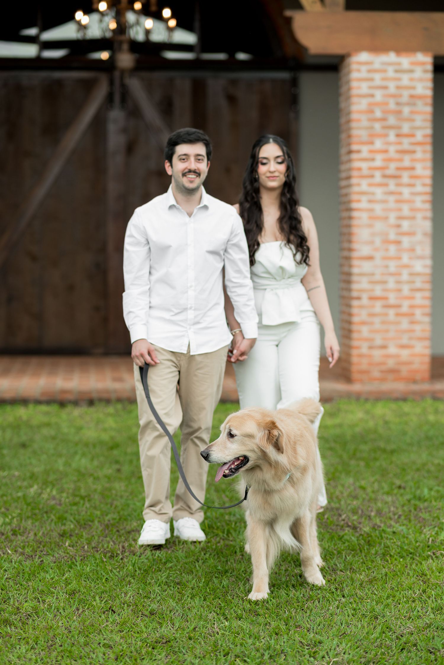 Smiling couple in white outfits walking a golden retriever on a leash on green grass near a brick building.