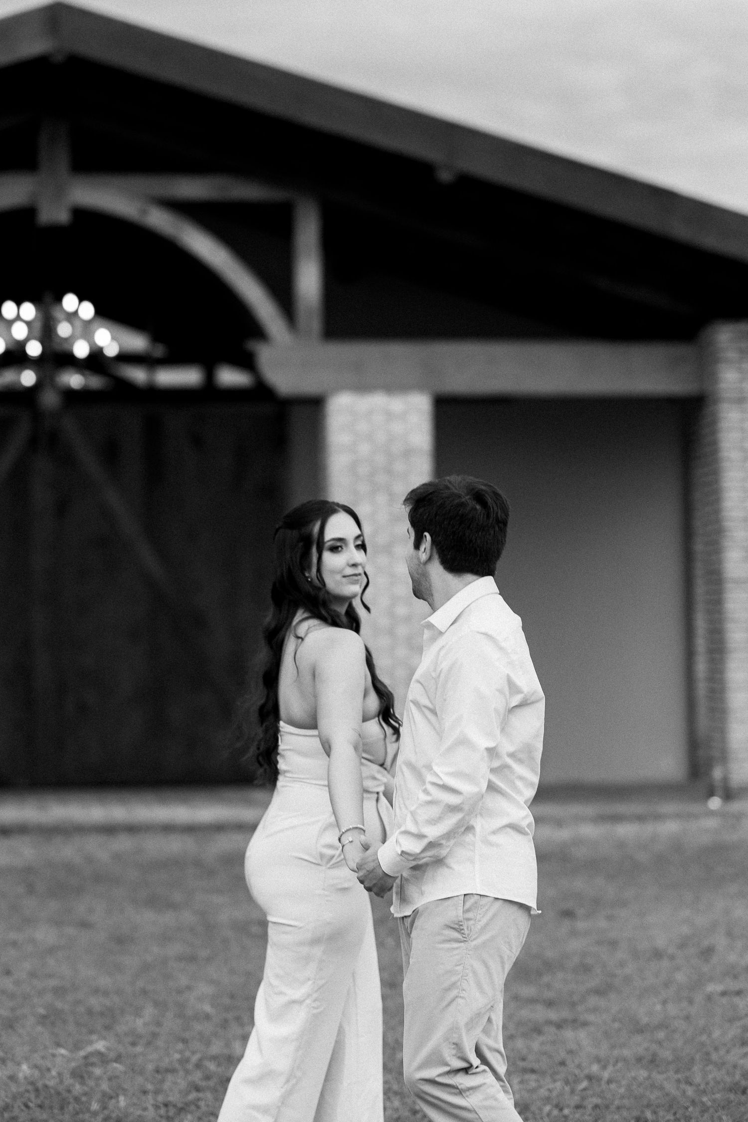 Black and white photo of a couple holding hands and smiling at each other in front of a rustic barn.