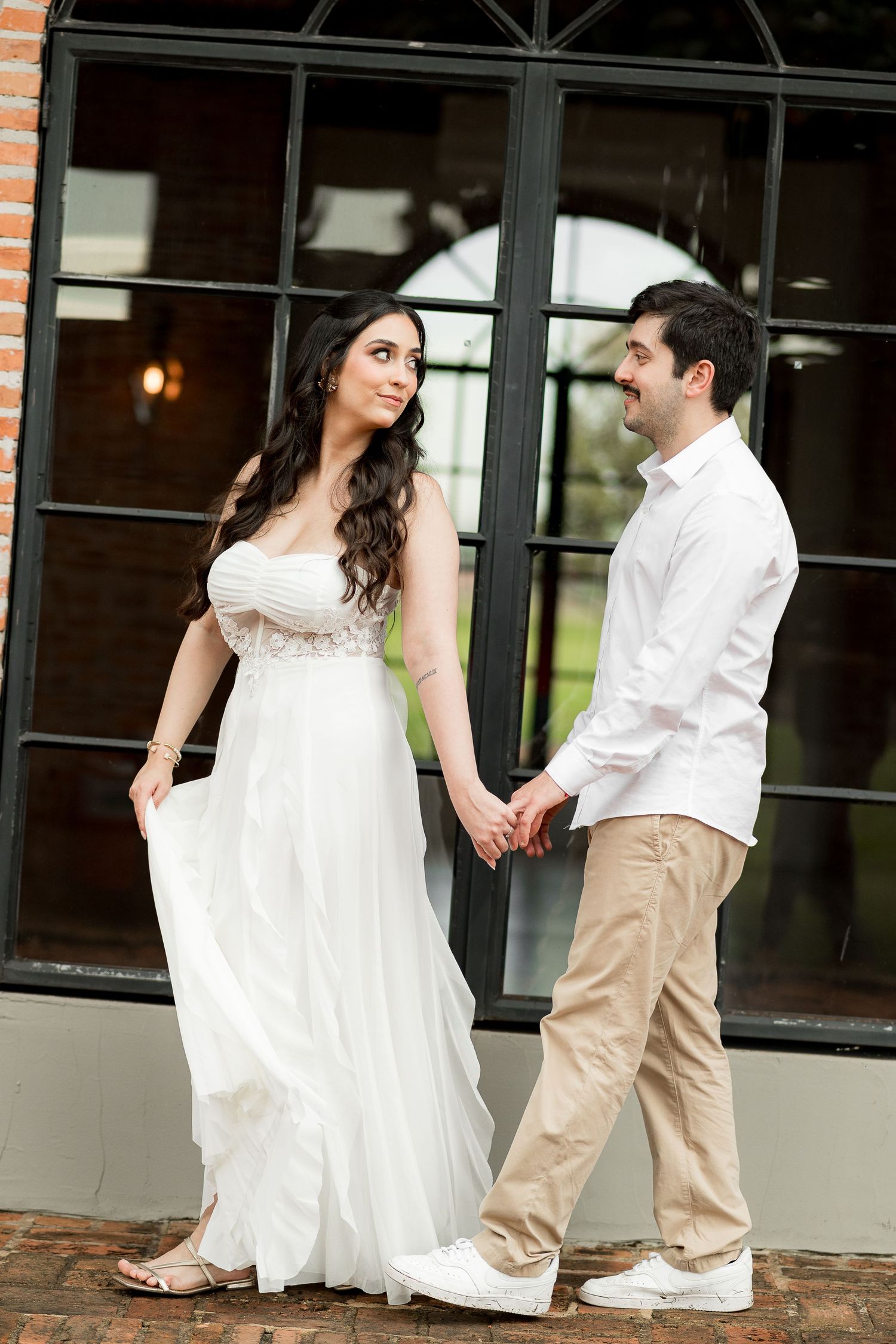 Couple holding hands in front of large black-framed window, woman in white flowing dress, man in white shirt and khaki pants.