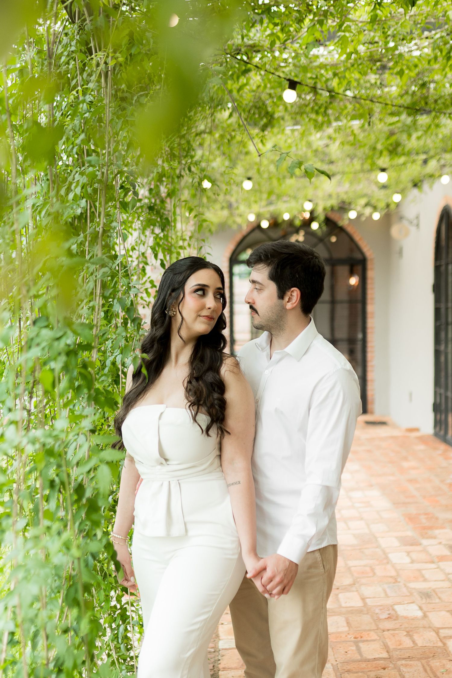 A couple in elegant white attire holds hands and smiles beneath a lush green vine-covered garden archway.