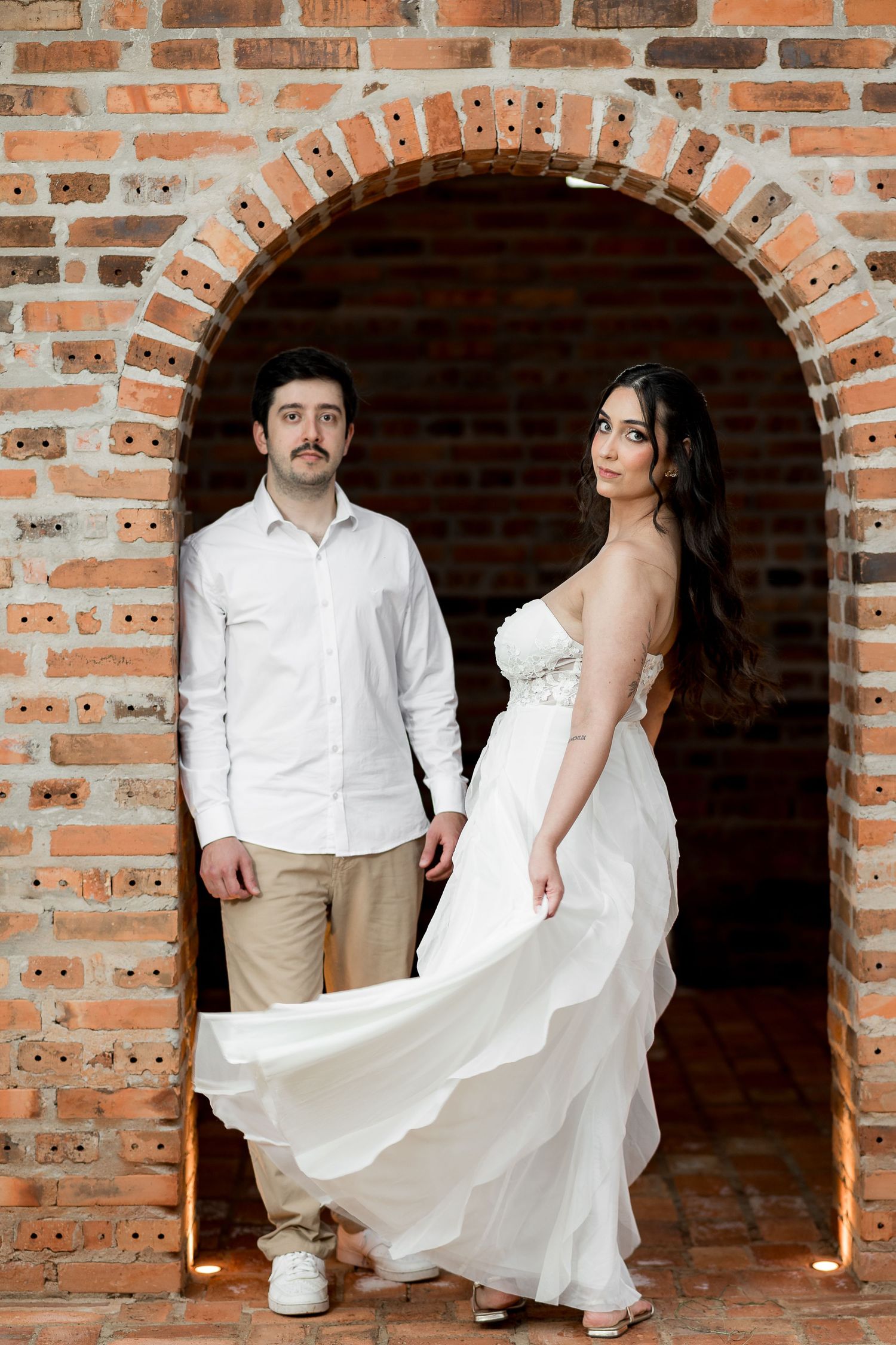 Couple posing in brick archway, man in white shirt and woman in flowing white strapless wedding gown.