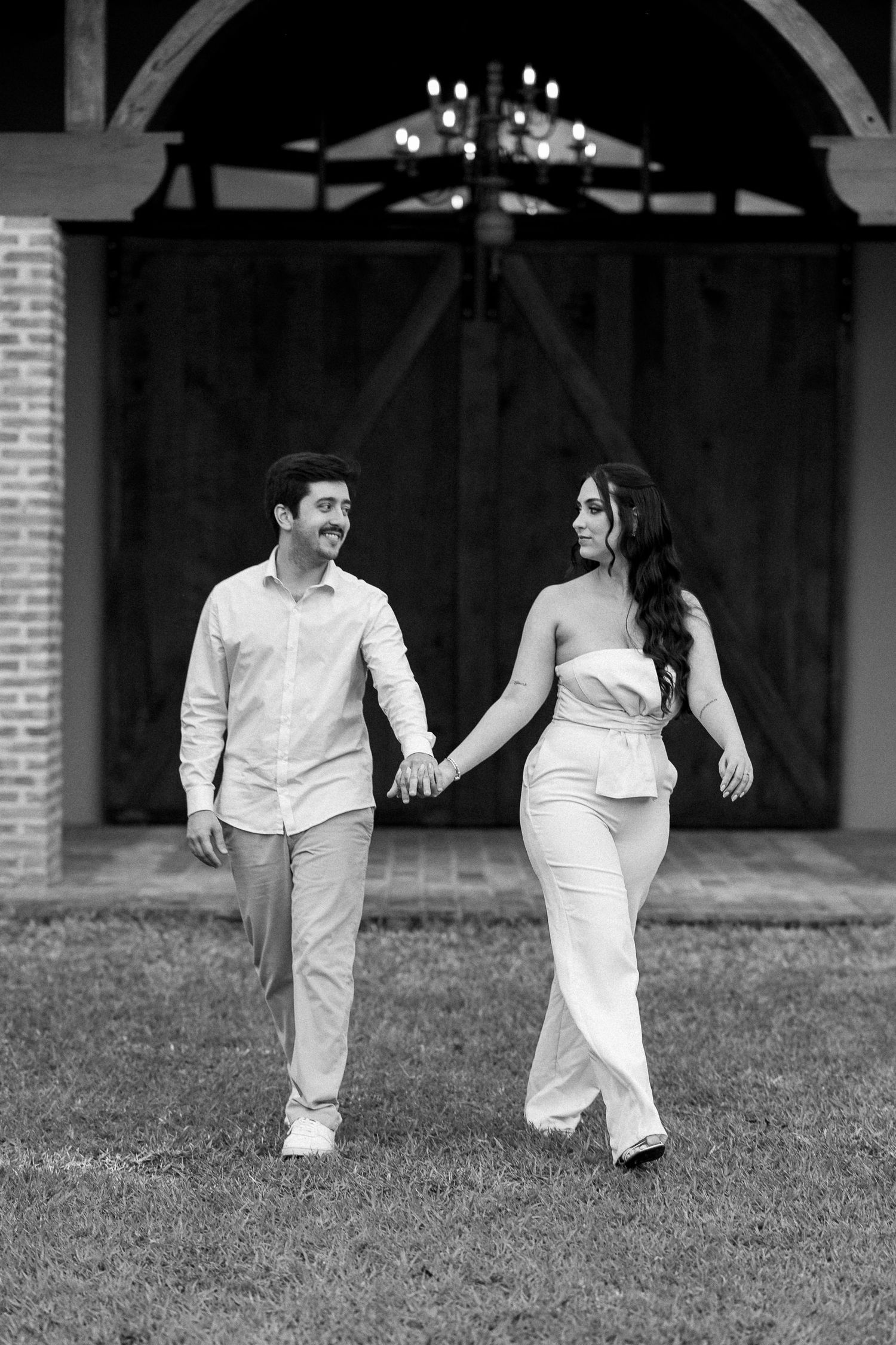 Black and white photo of a couple holding hands and walking in front of a rustic barn with a chandelier.