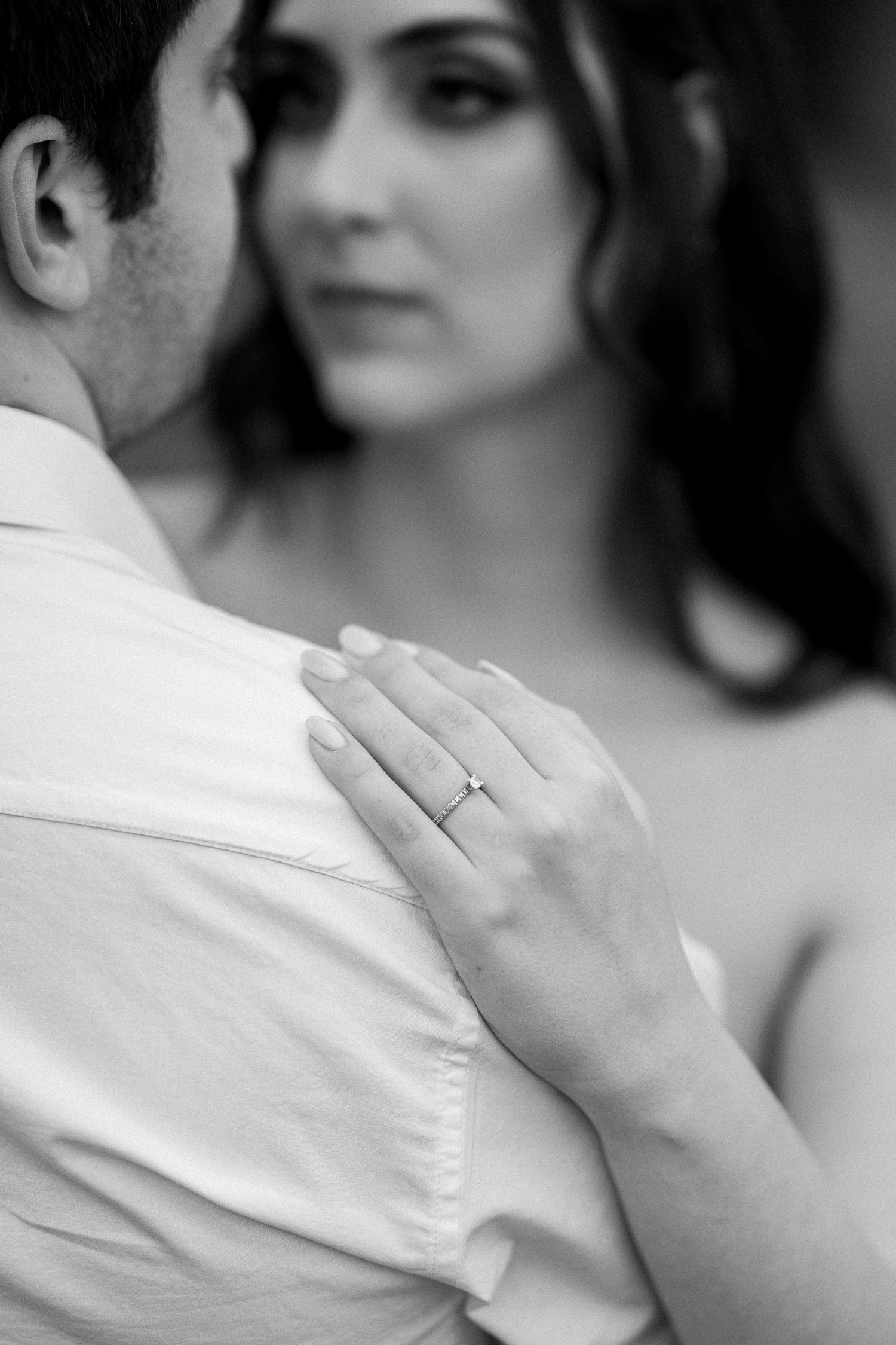 Black and white photo of a couple embracing, woman's hand resting on man's shoulder displaying an elegant engagement ring.