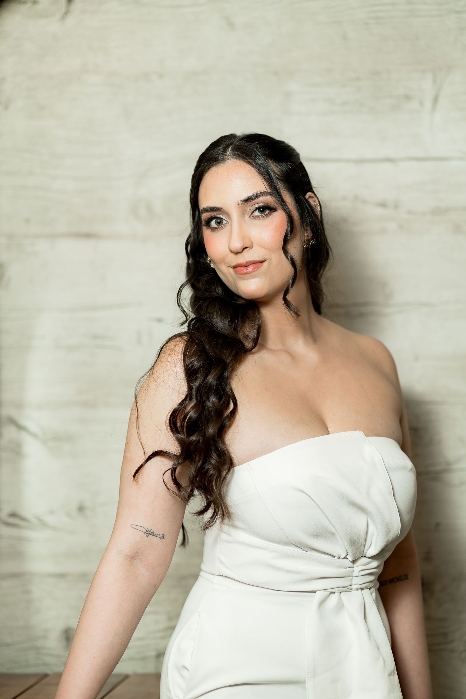 Woman in white strapless gown with wavy dark hair and subtle makeup poses elegantly against a light textured wall.
