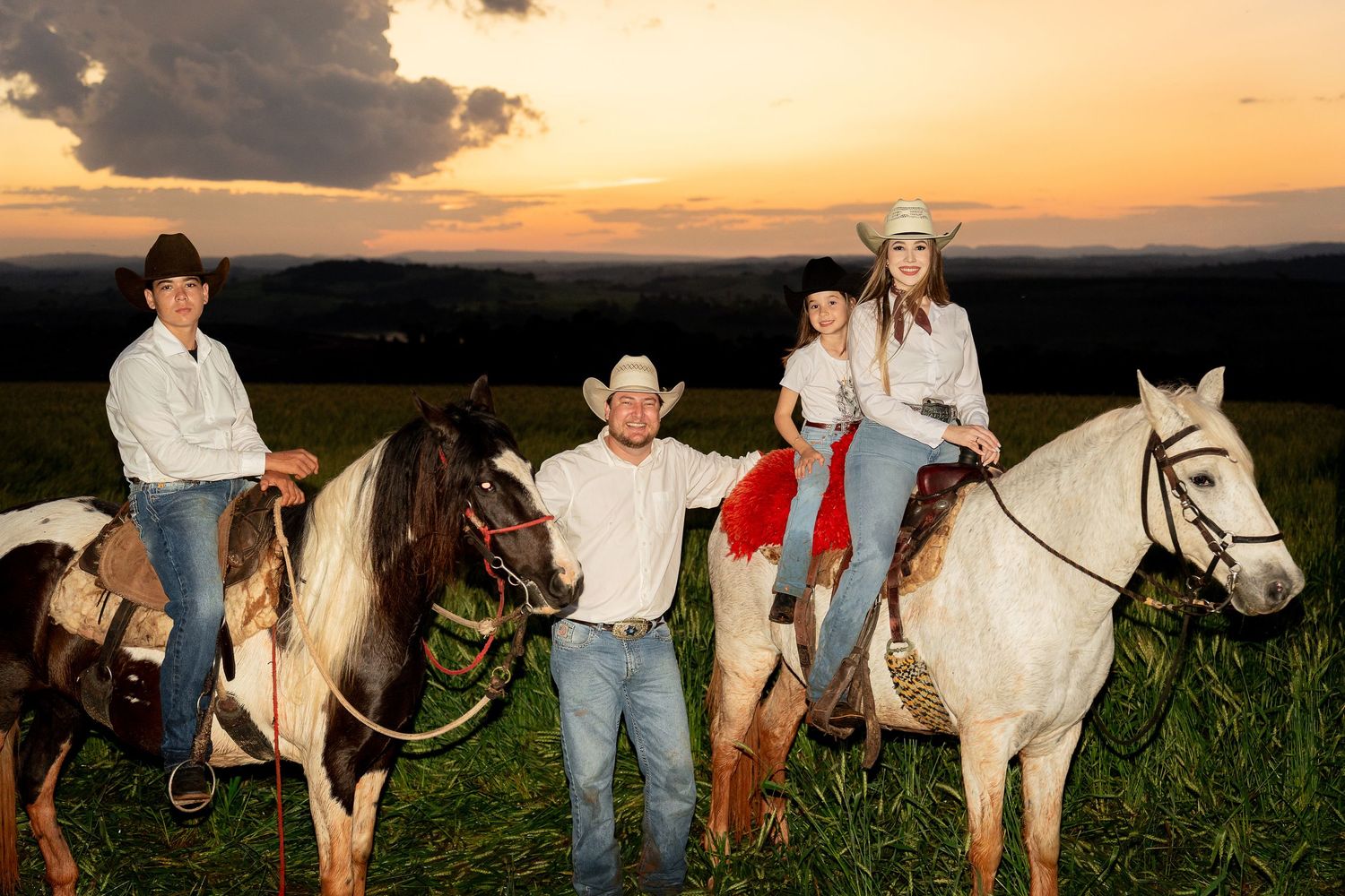 Family of four in cowboy hats posing on horseback at sunset in a green field.