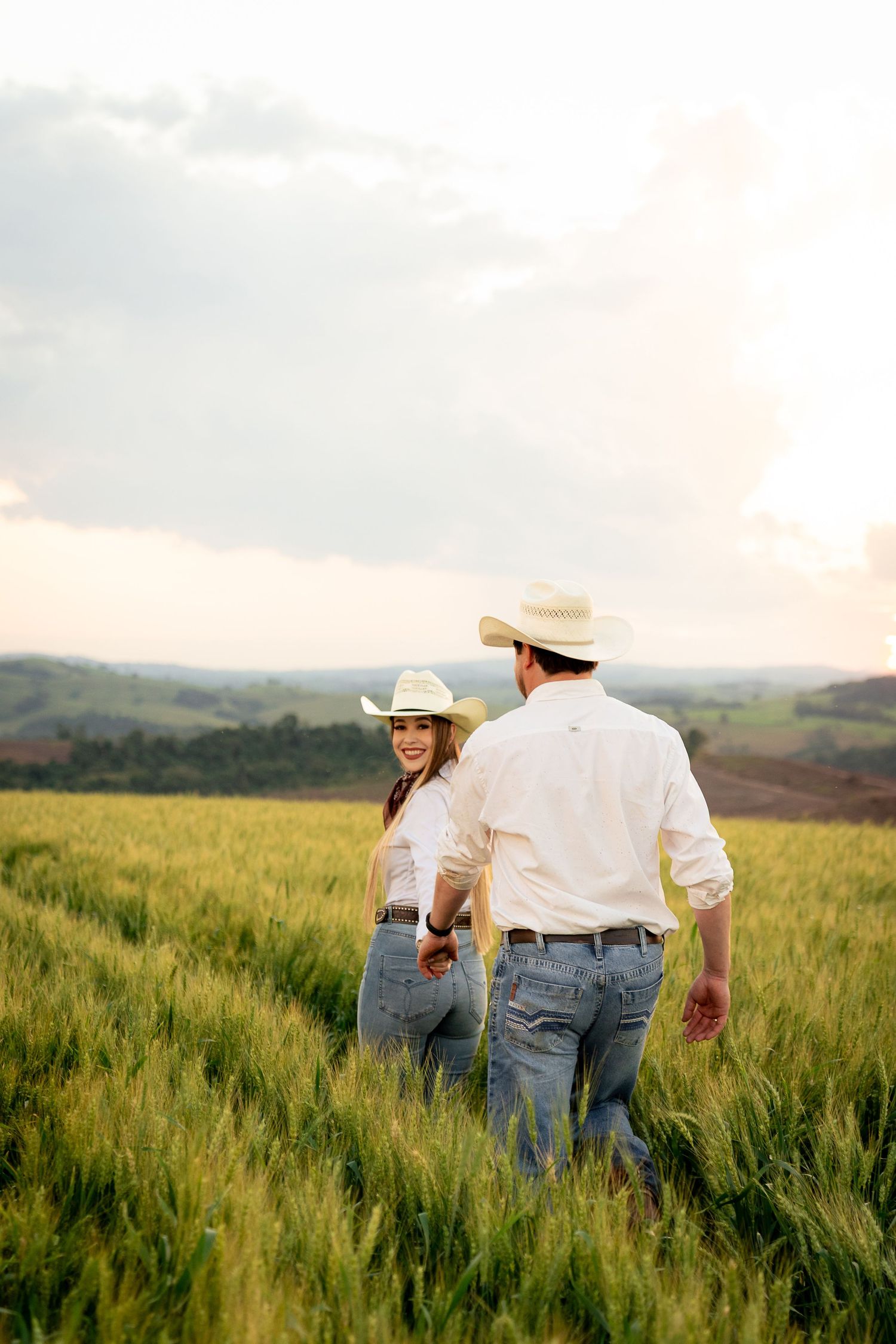 A couple in cowboy hats and jeans hold hands while walking through a lush green wheat field at sunset.