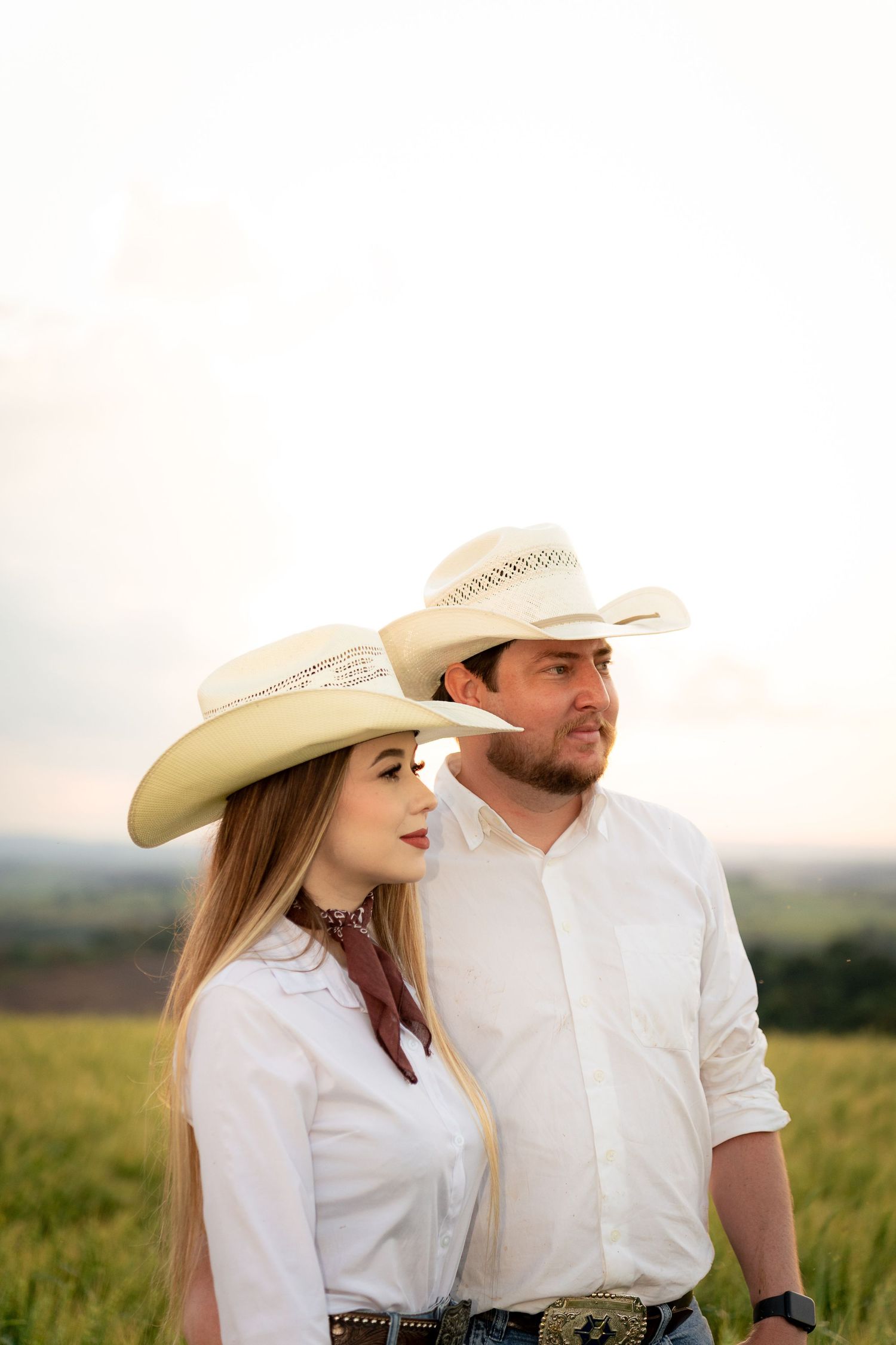 A couple in matching white cowboy hats and shirts stand together in a green field at sunset.