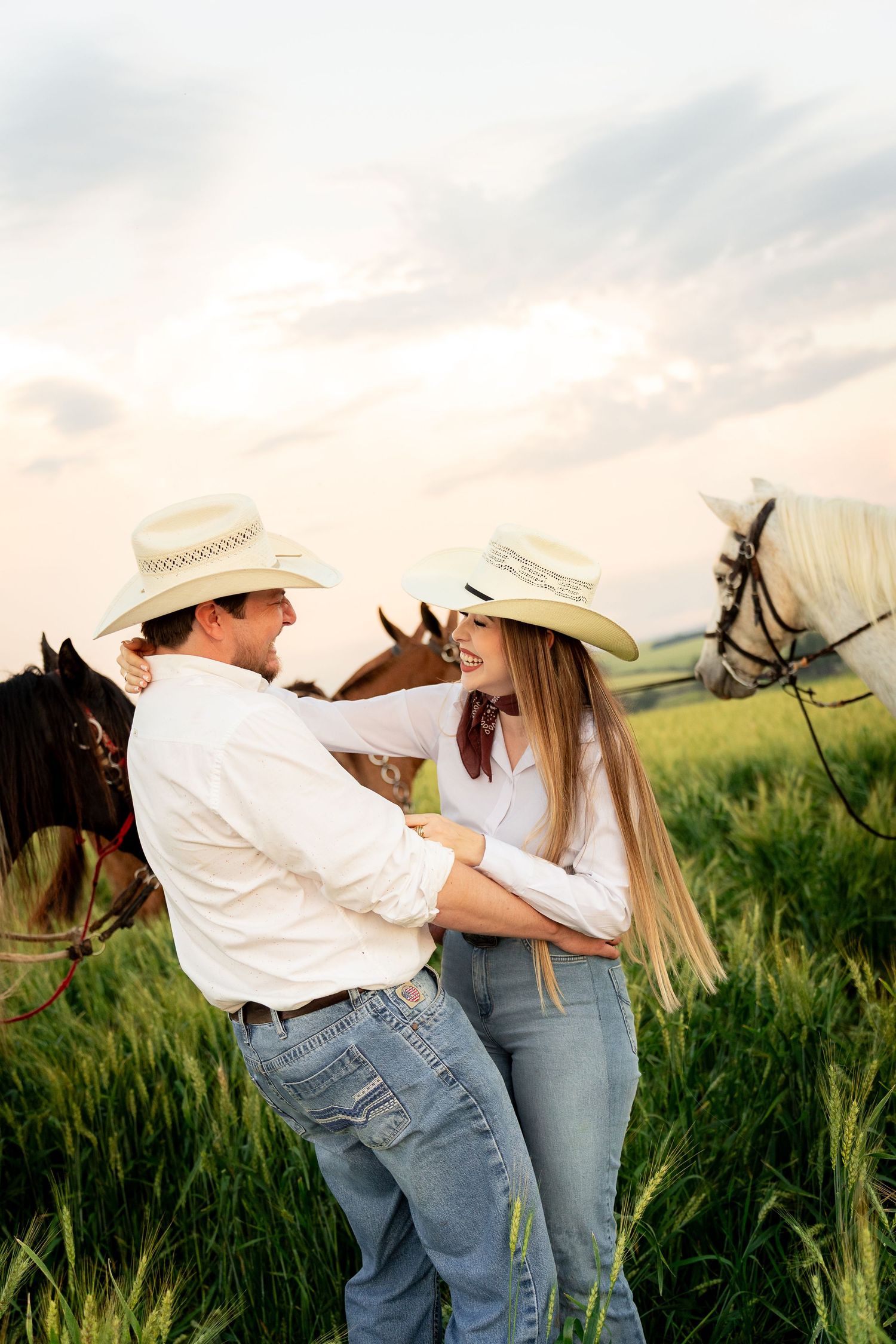 A couple in cowboy hats and jeans embrace in a green field with horses at sunset.