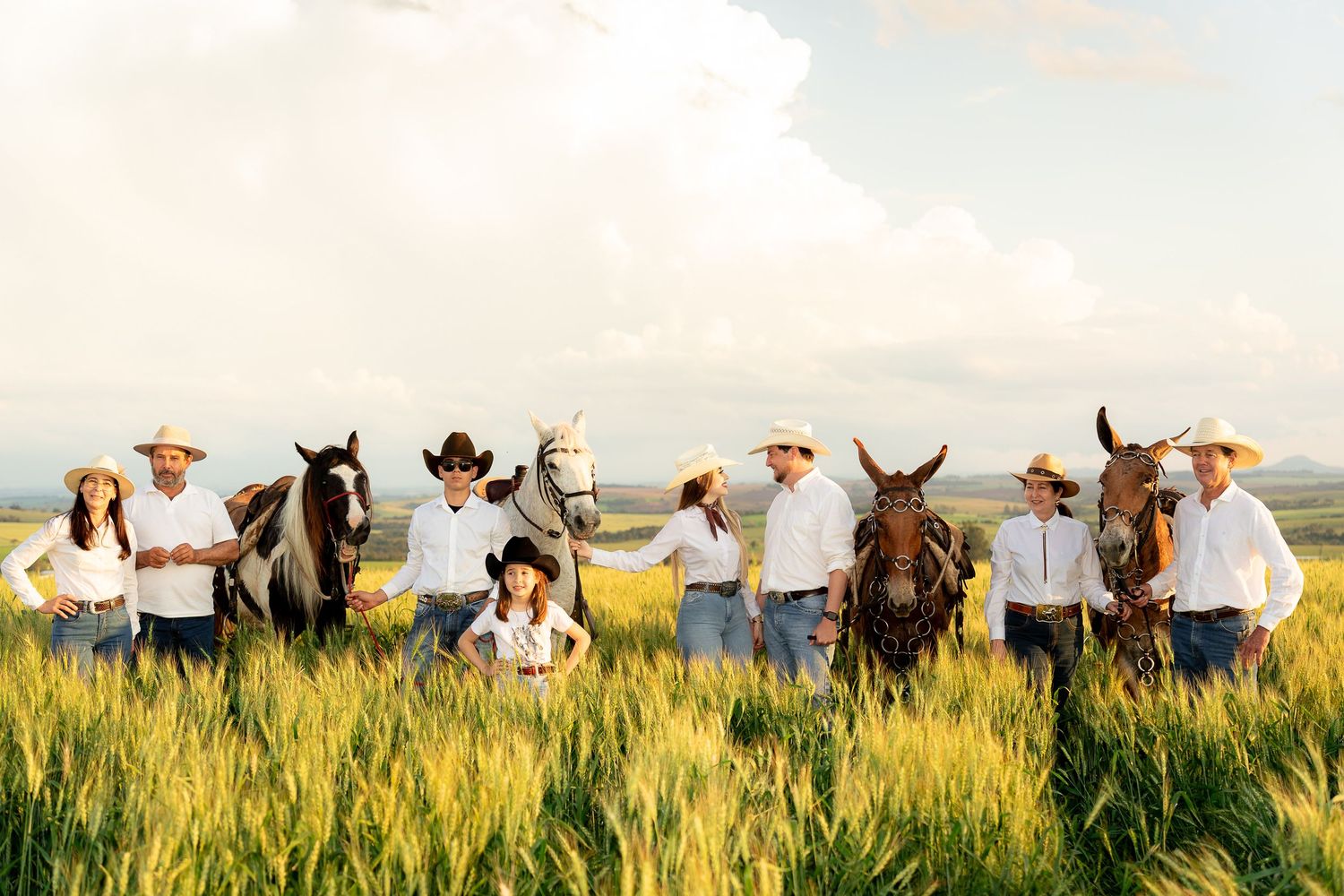 A family of cowboys and cowgirls pose with horses in a lush green field under a dramatic cloudy sky.