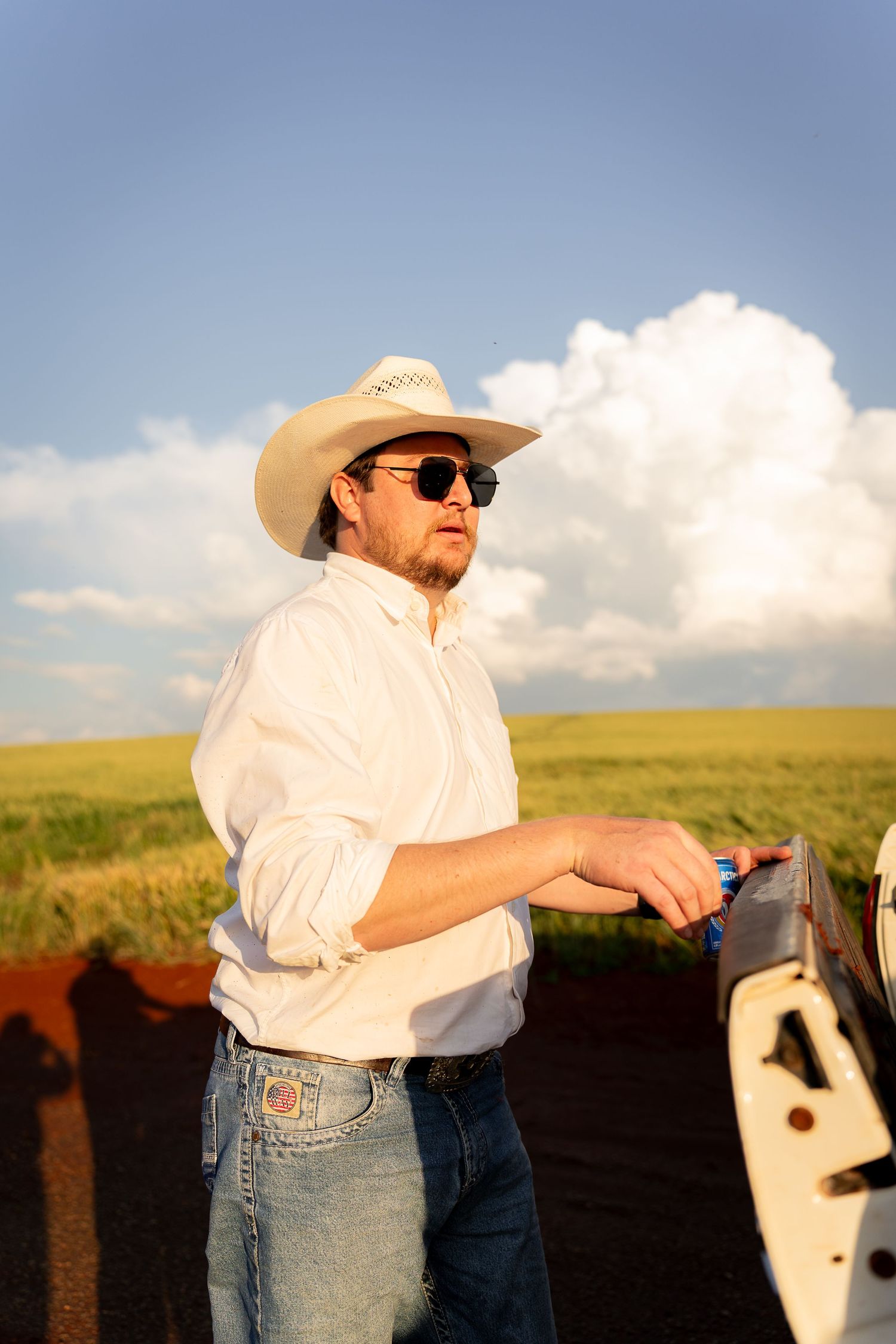 A cowboy in a white hat, sunglasses, and jeans stands by a truck in a green field under a blue sky.
