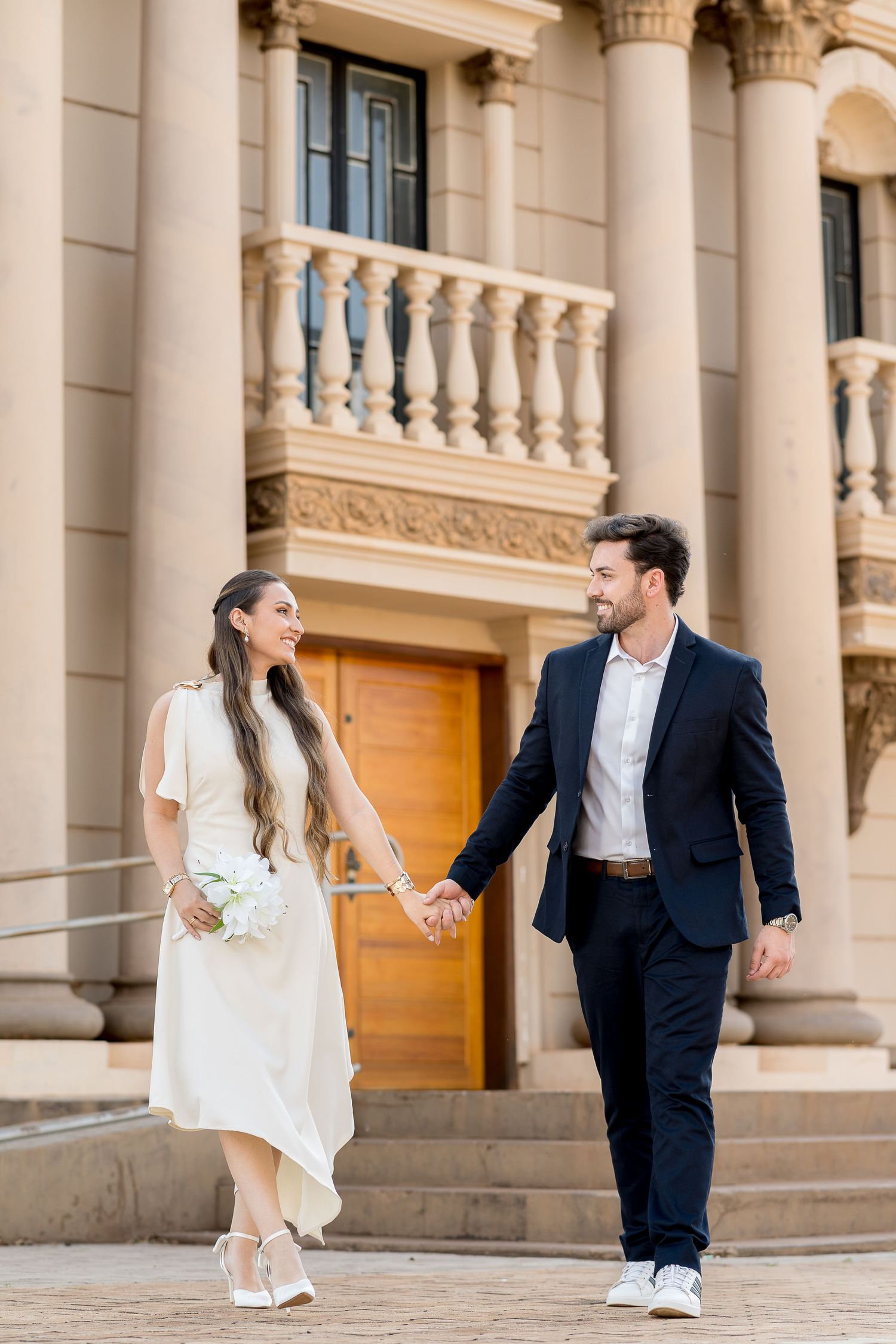 A smiling bride and groom hold hands walking in front of a classical building, she carries white flowers in a white dress.